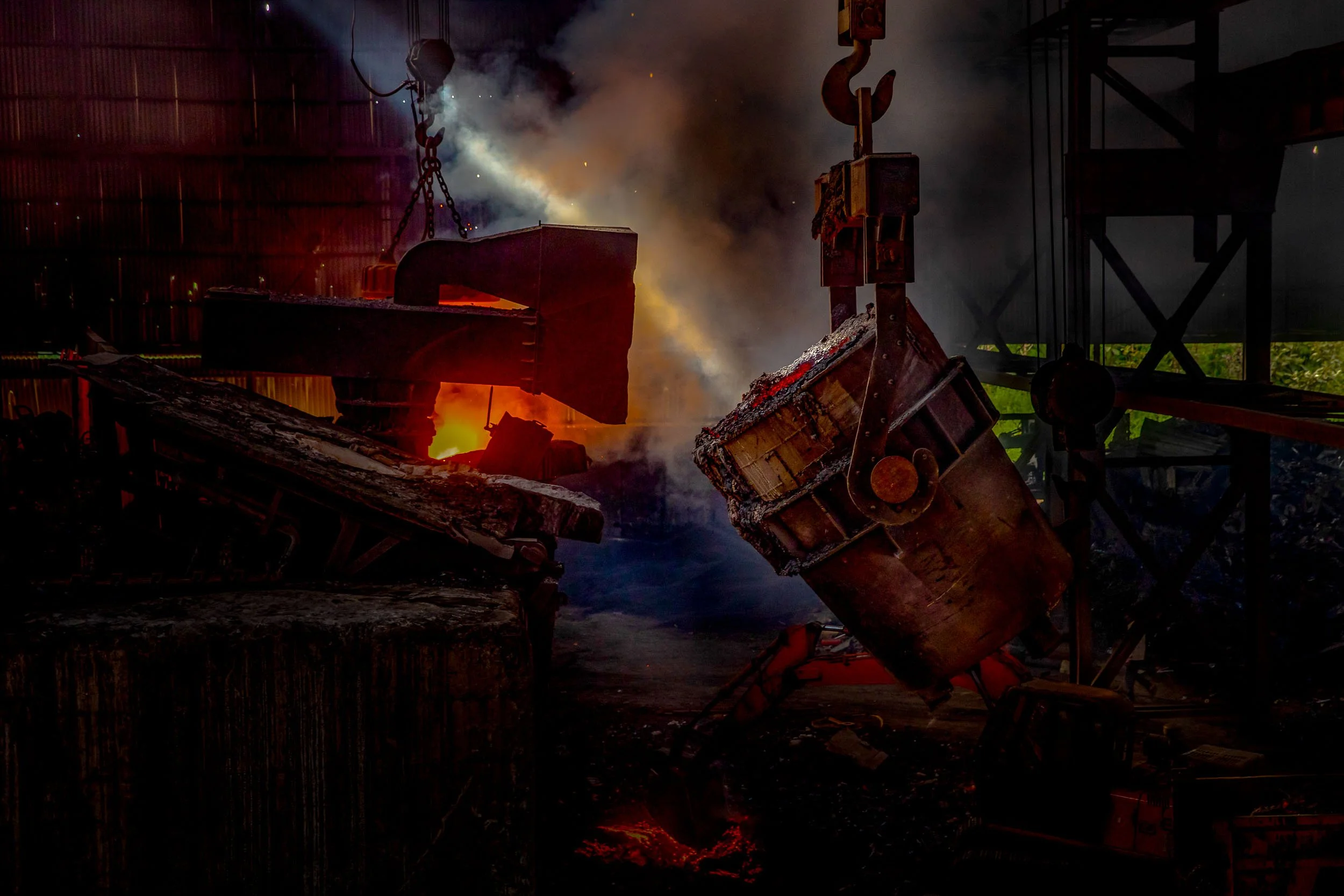 Interior industrial photography of a massive ladle pouring glowing molten metal in a heavy manufacturing steel foundry.