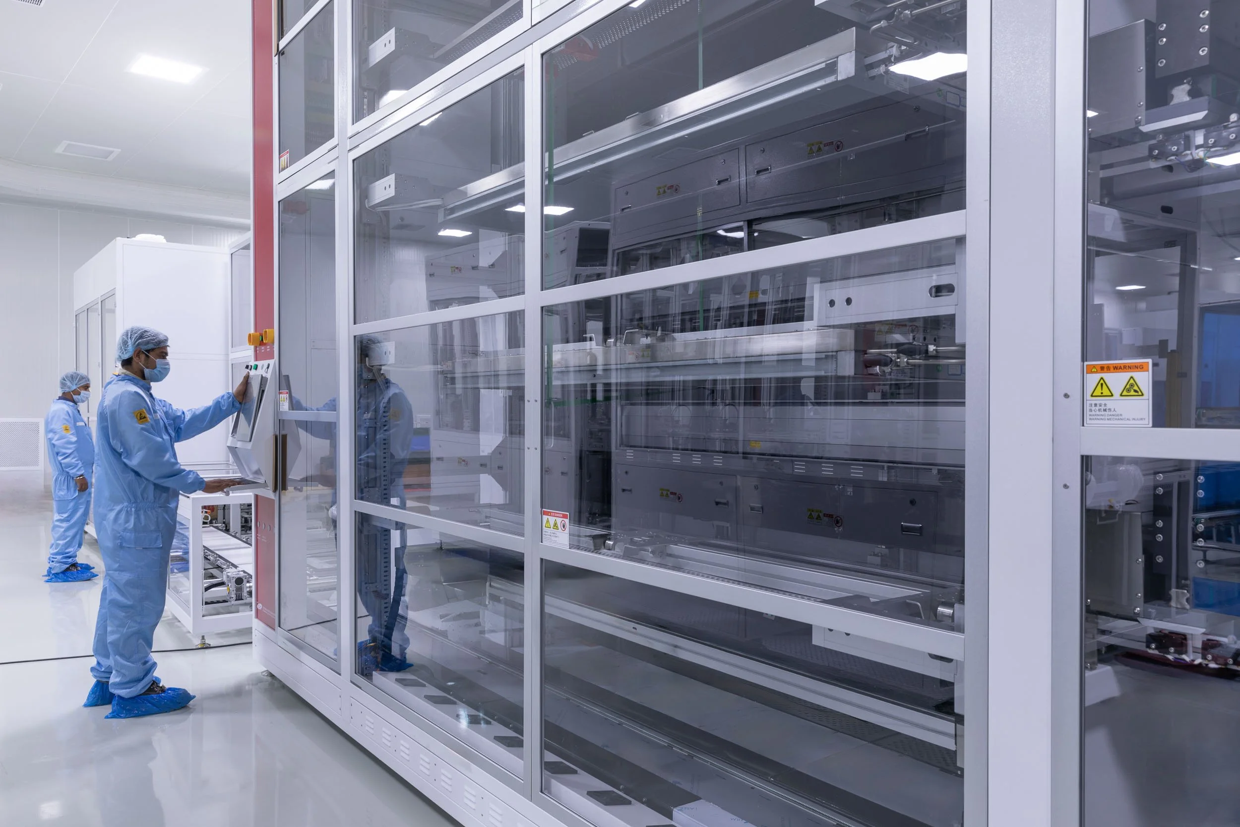 Interior industrial photography of workers in blue cleanroom suits operating an enclosed glass machinery unit in a sterile advanced manufacturing facility