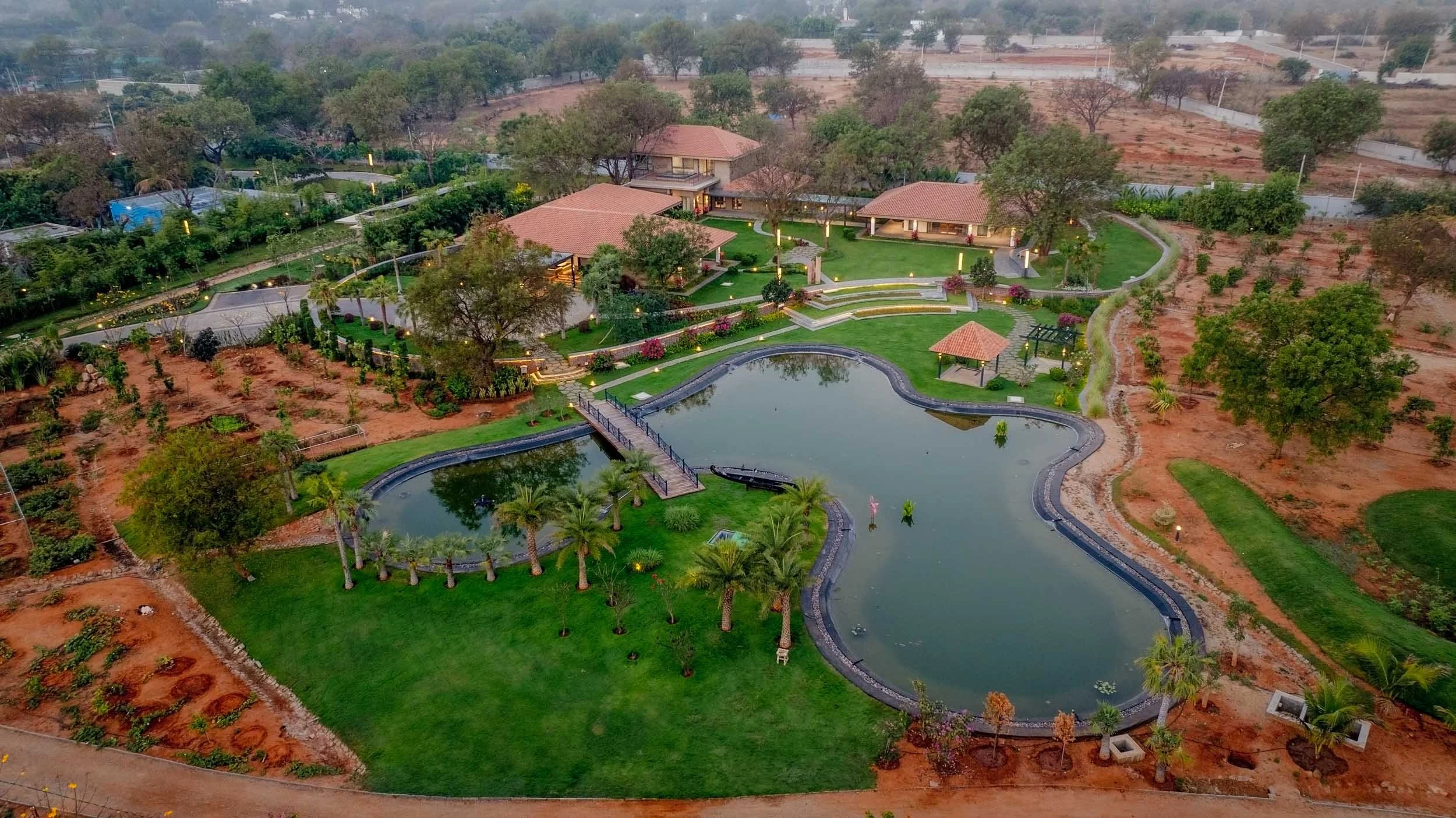 Aerial landscape photography of a sprawling resort estate featuring a large organic pond, lush green lawns, and terracotta-roofed villas