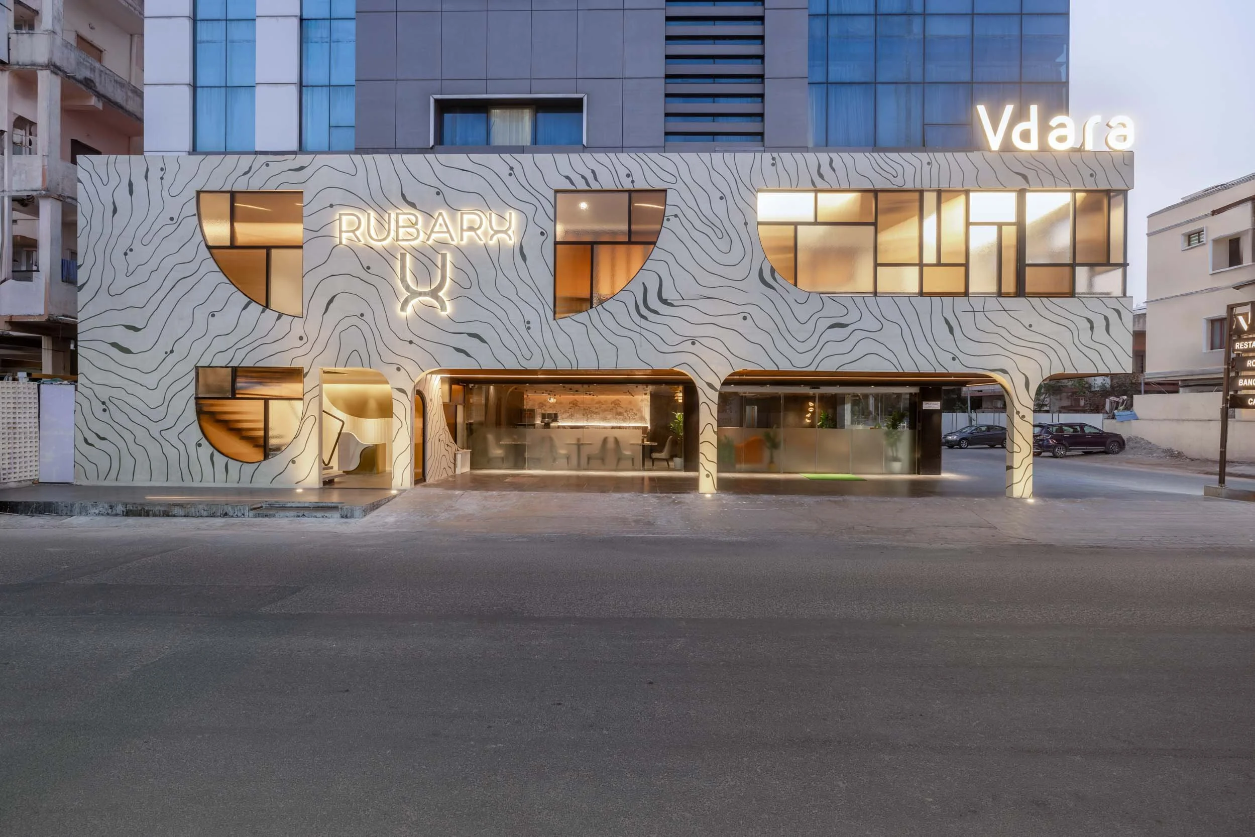 An exterior twilight view of a modern commercial building, featuring a distinctive textured white facade with topographical line patterns, large arched entrances, and warm interior illumination