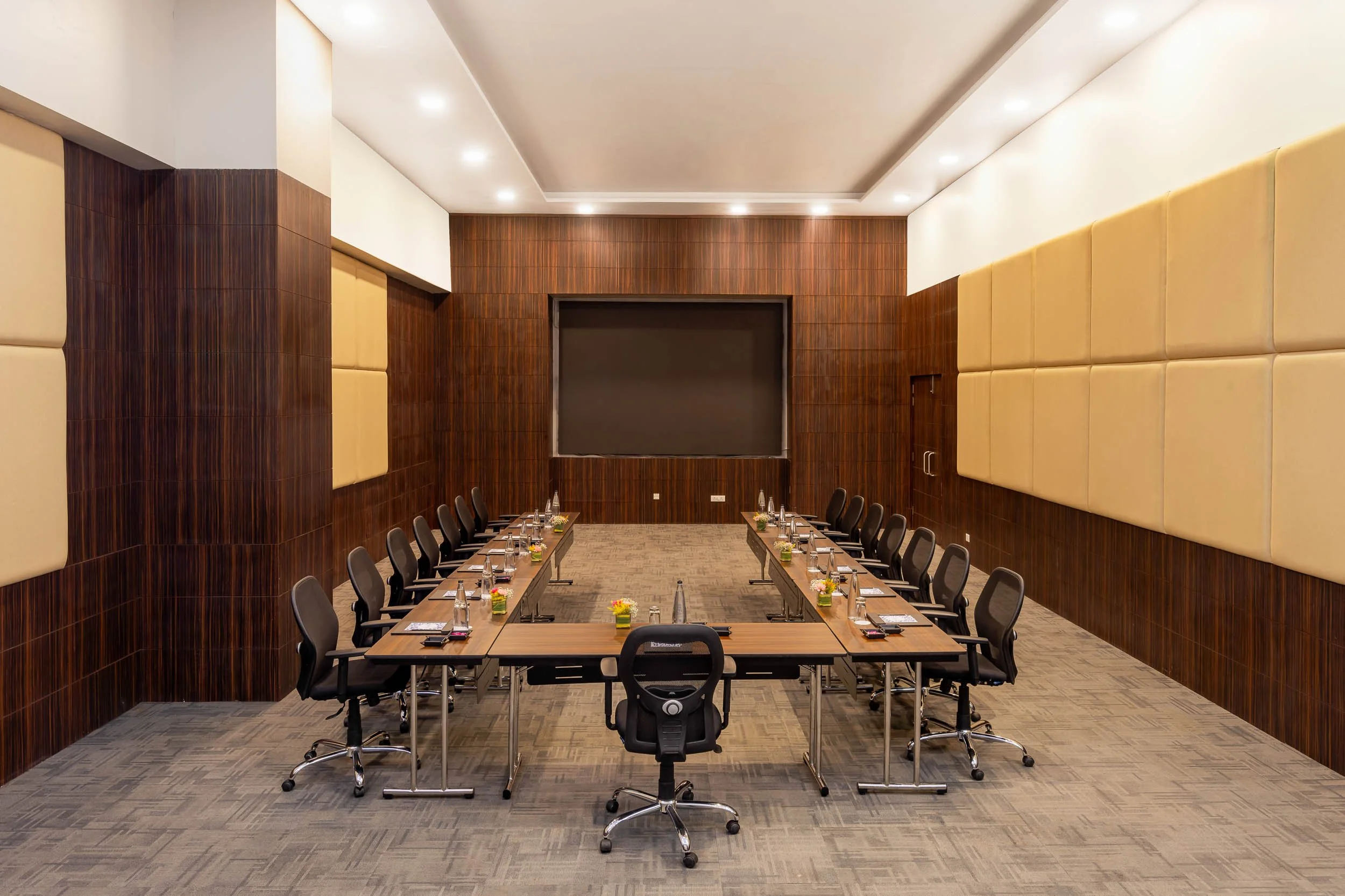 Interior photography of a modern hotel meeting room featuring a long wooden conference table, black ergonomic chairs, wood-paneled walls, and a large presentation screen