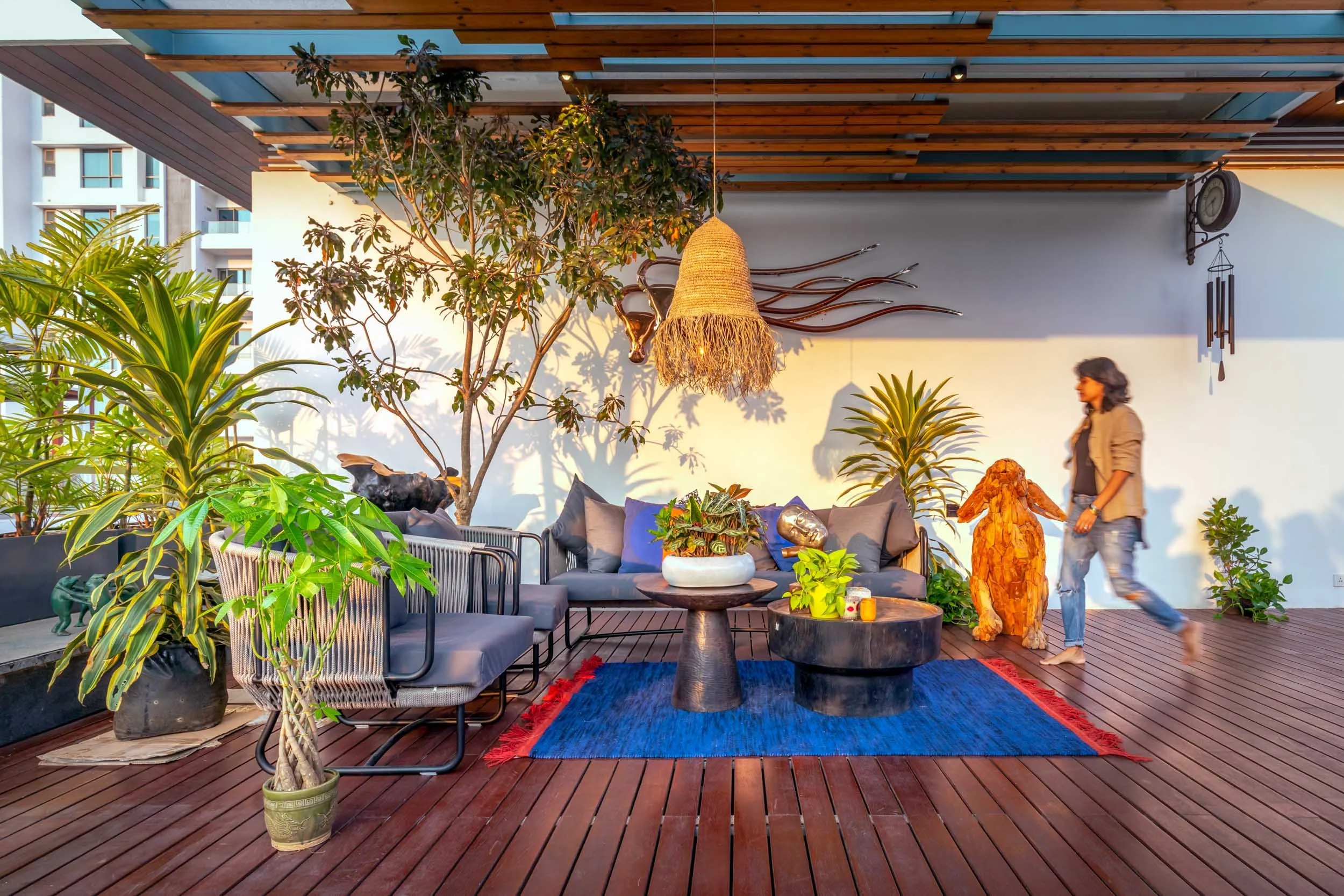 An outdoor living space featuring wooden decking, a pergola, comfortable seating on a blue rug, and surrounded by lush green plants