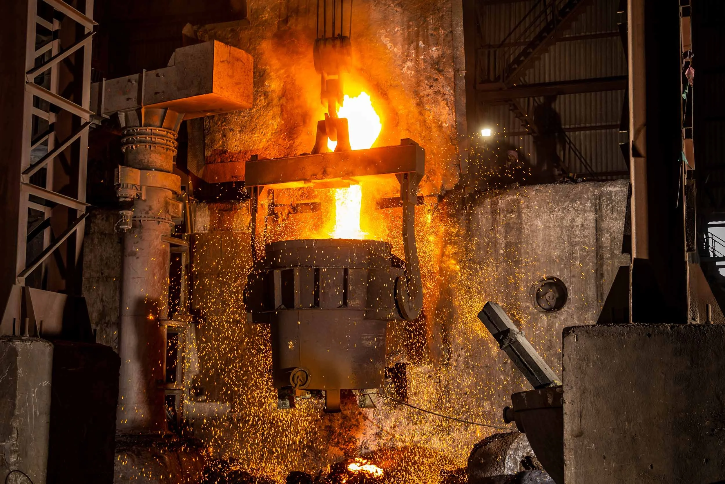 Interior industrial photography of a massive ladle pouring glowing molten metal in a heavy manufacturing steel foundry.