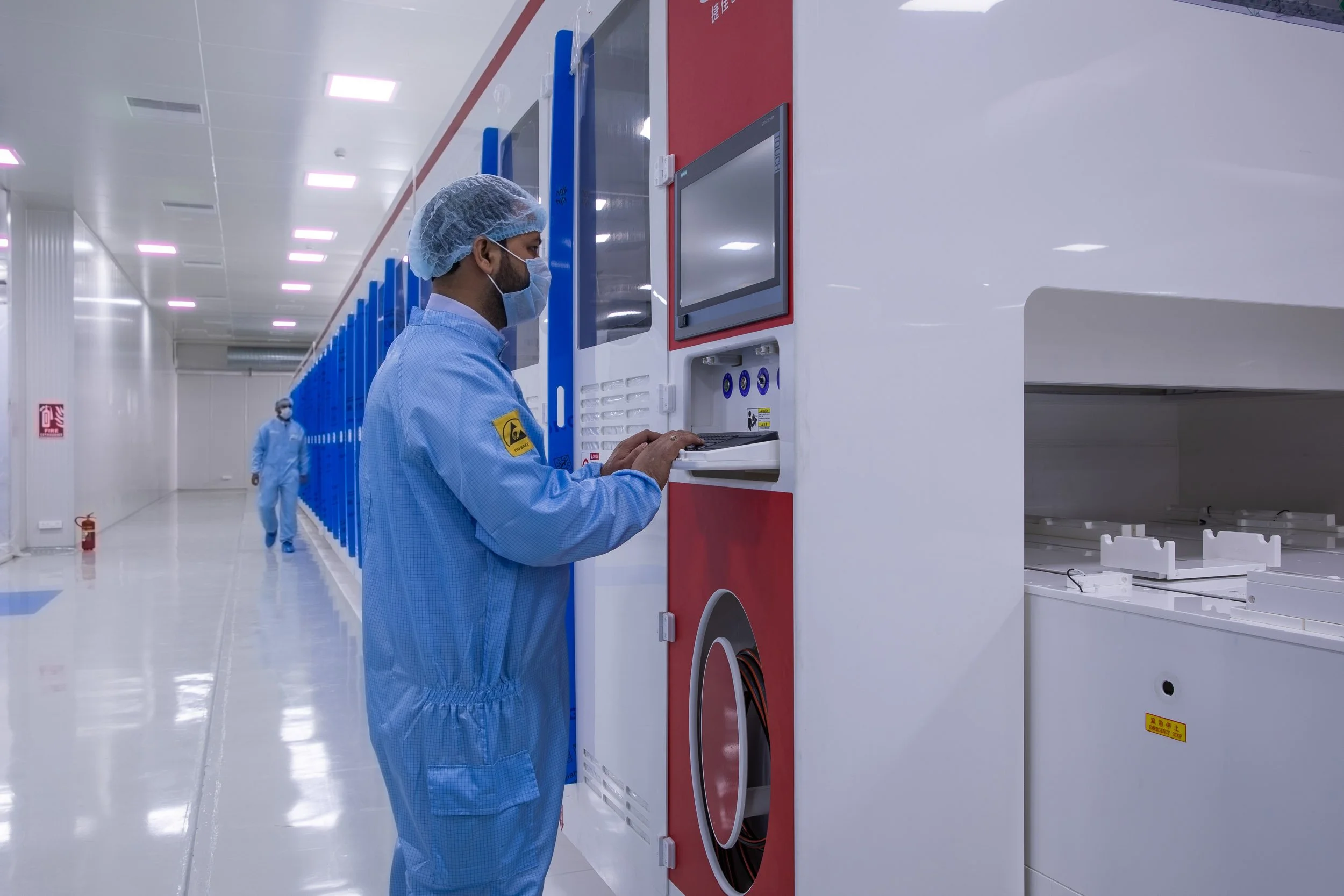 Interior industrial photography of a worker in a blue cleanroom suit operating a specialized blue and red machine in a sterile manufacturing facility.