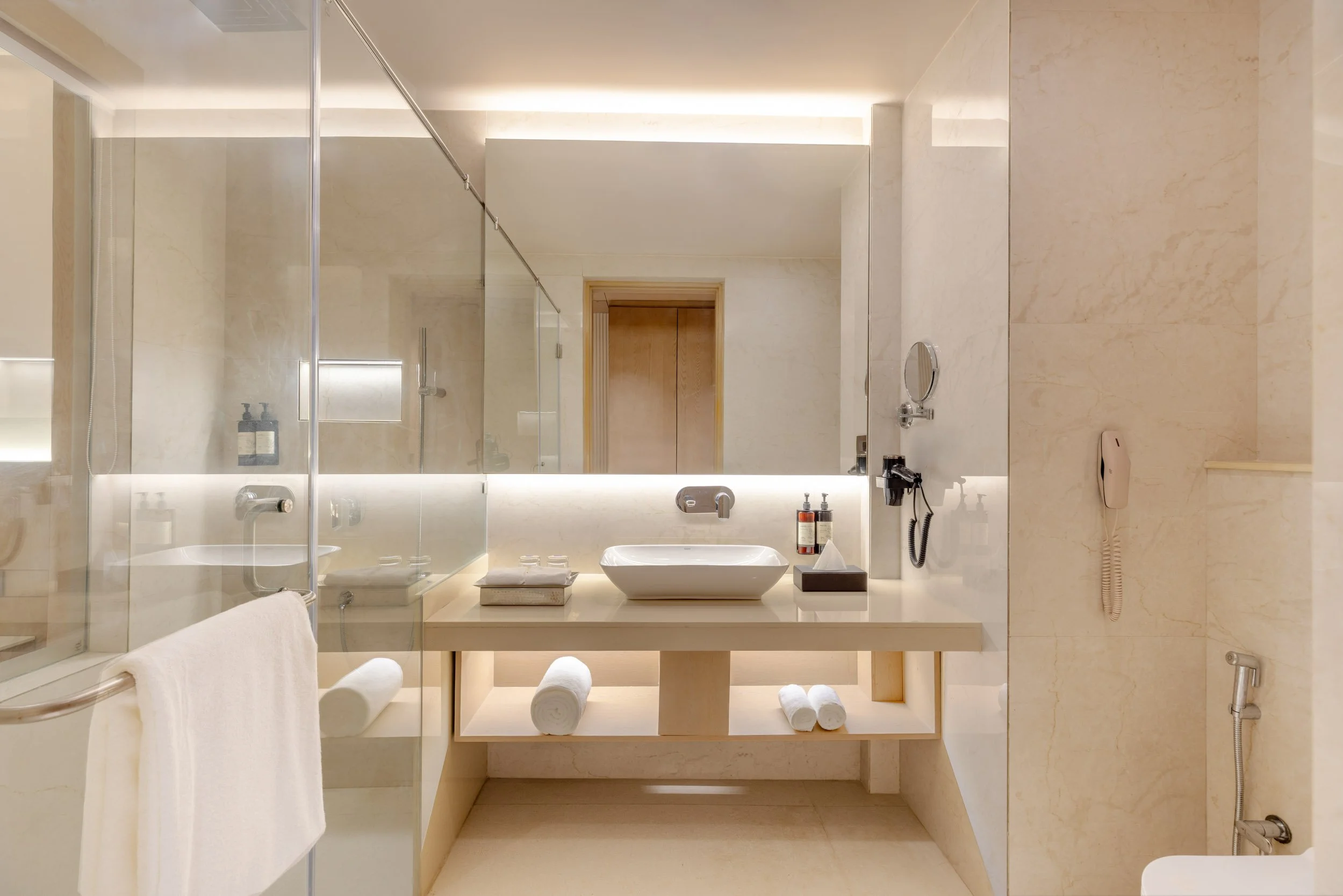 Interior photography of a luxury hotel bathroom featuring a large glass-enclosed shower, a wide vanity with an expansive mirror, and warm beige wall tiles