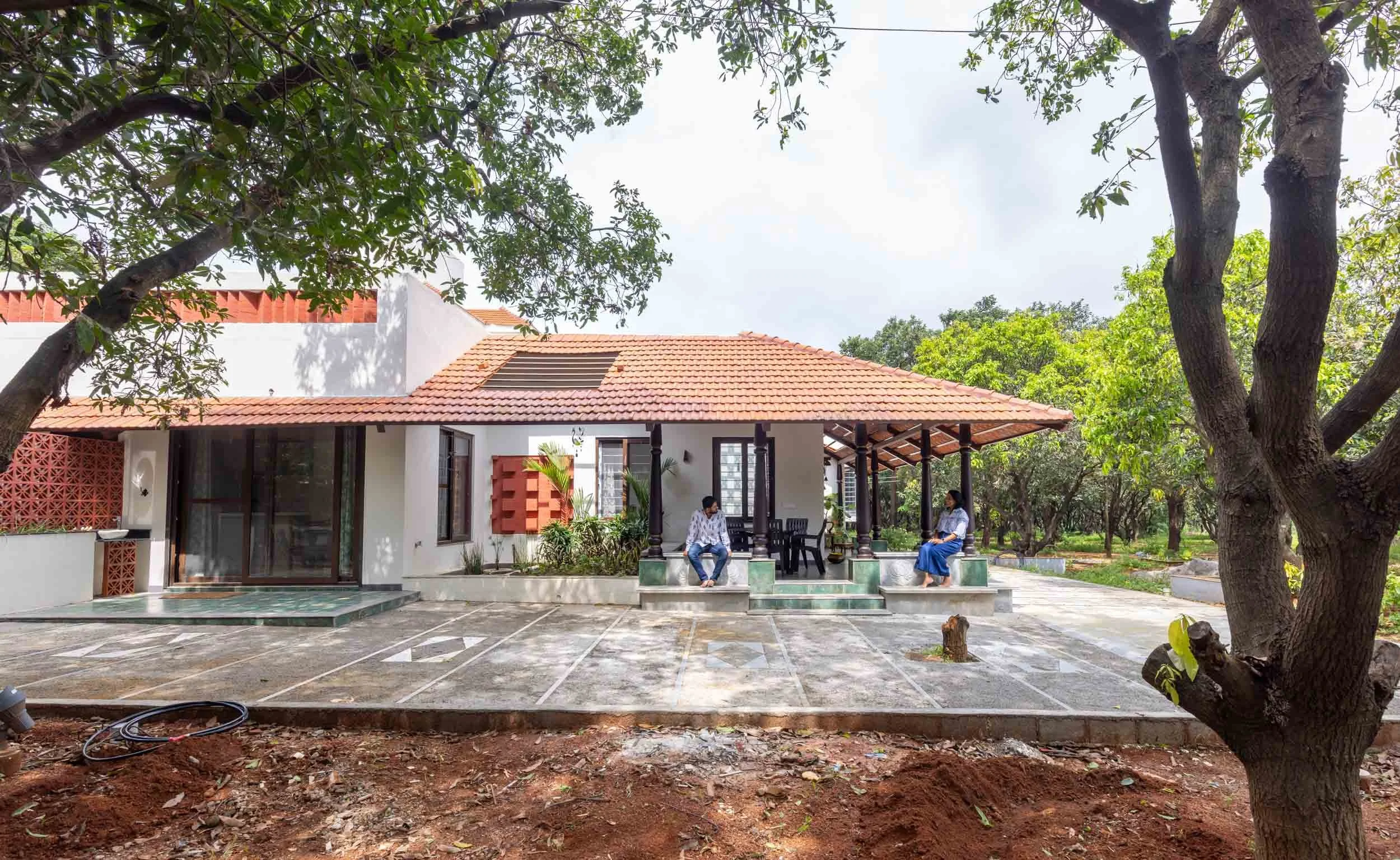 Professional exterior architectural photography of a sprawling residential farmhouse, highlighting its traditional terracotta tiled roof, wide veranda, and lush green landscape.