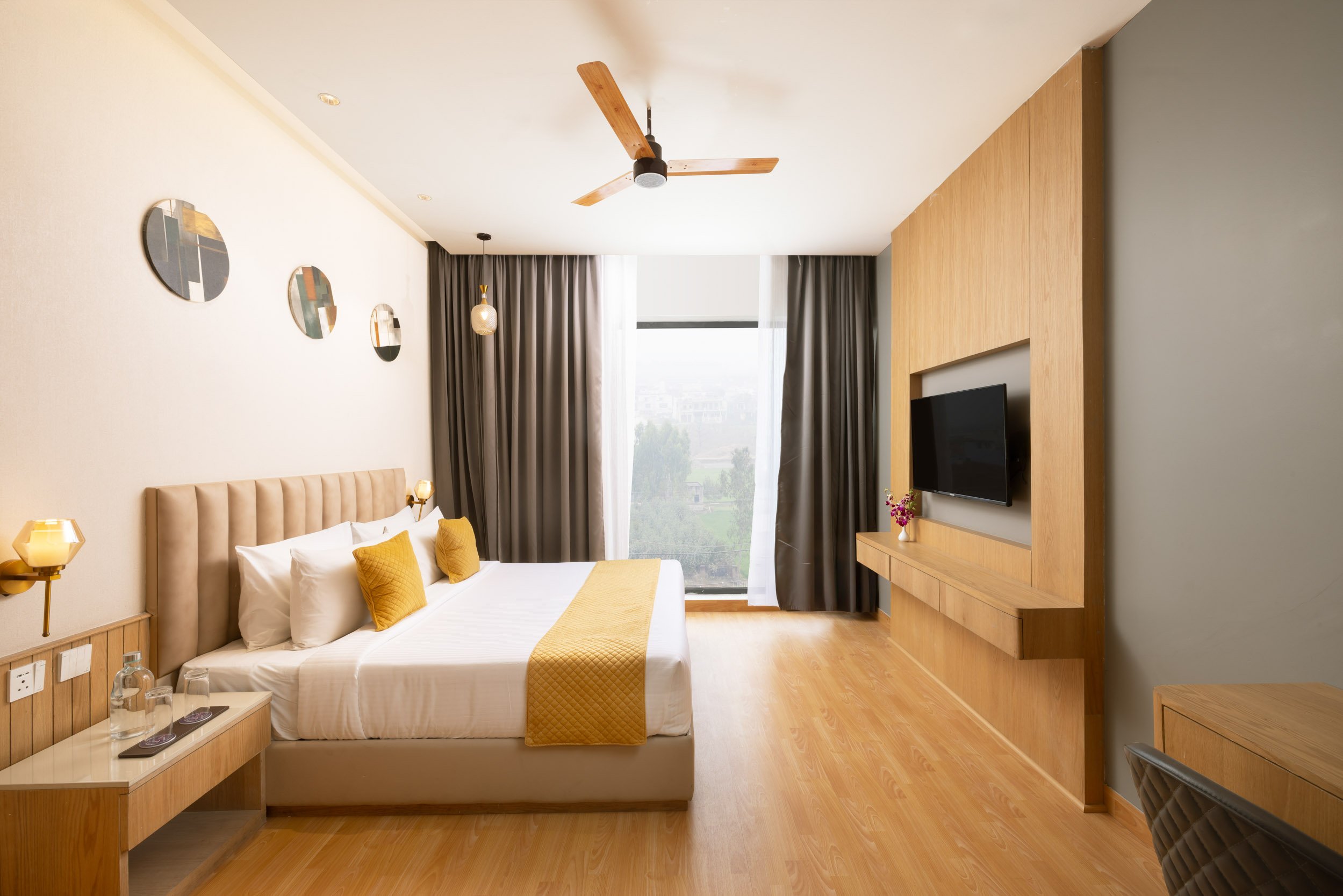Interior photography of a modern hotel king room featuring a bed with a yellow runner, wooden flooring, a wall-mounted television, and a large window.