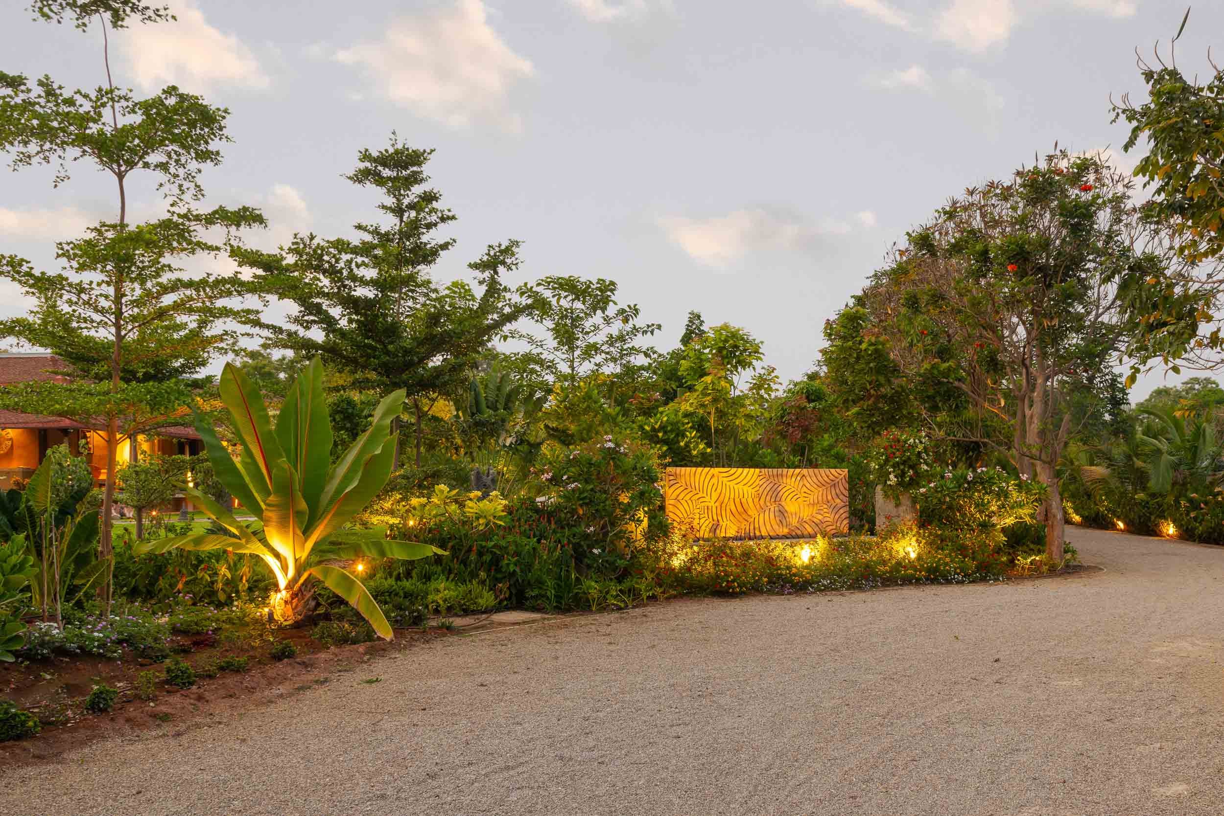 Exterior twilight photography of a luxury resort farm stay entrance featuring an illuminated stone wall, a wide driveway, and mature trees against an evening sky.