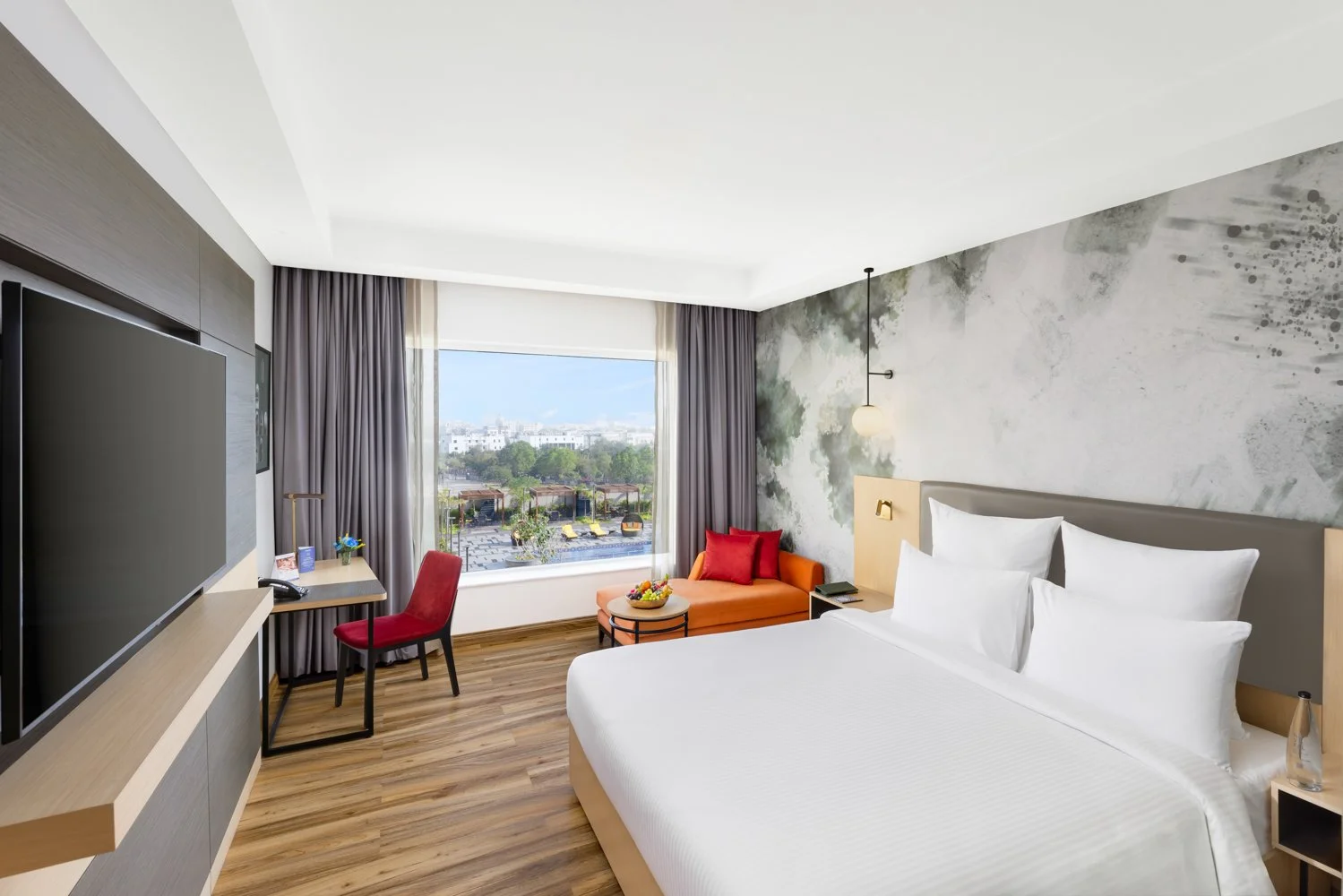 Wide interior photography of a modern hotel bedroom featuring a large bed with a custom nature mural, a wall-mounted TV, wooden flooring, and an orange armchair by the window