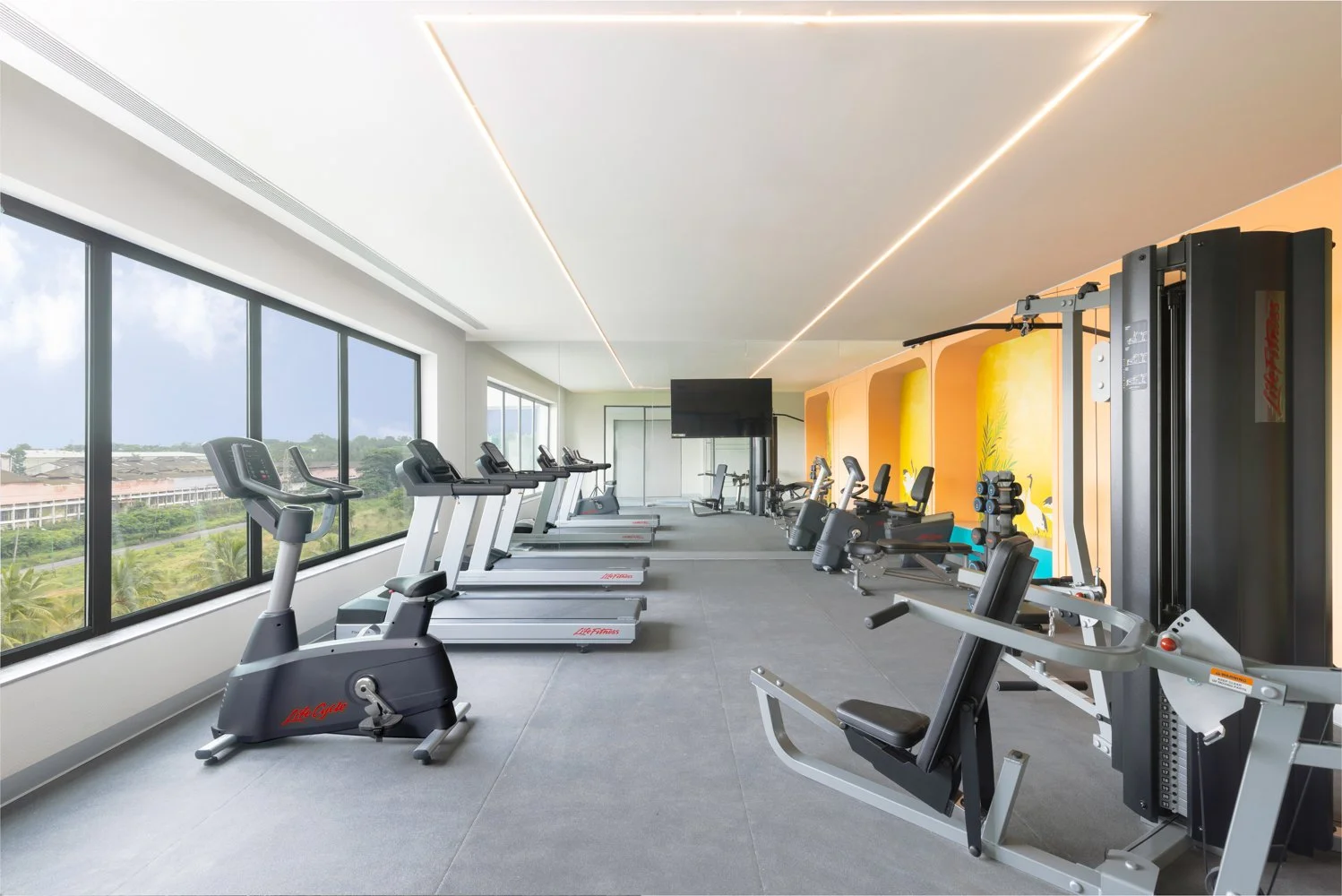 Interior photography of a modern hotel gym featuring rows of treadmills and ellipticals, large floor-to-ceiling windows, and bright yellow accent walls