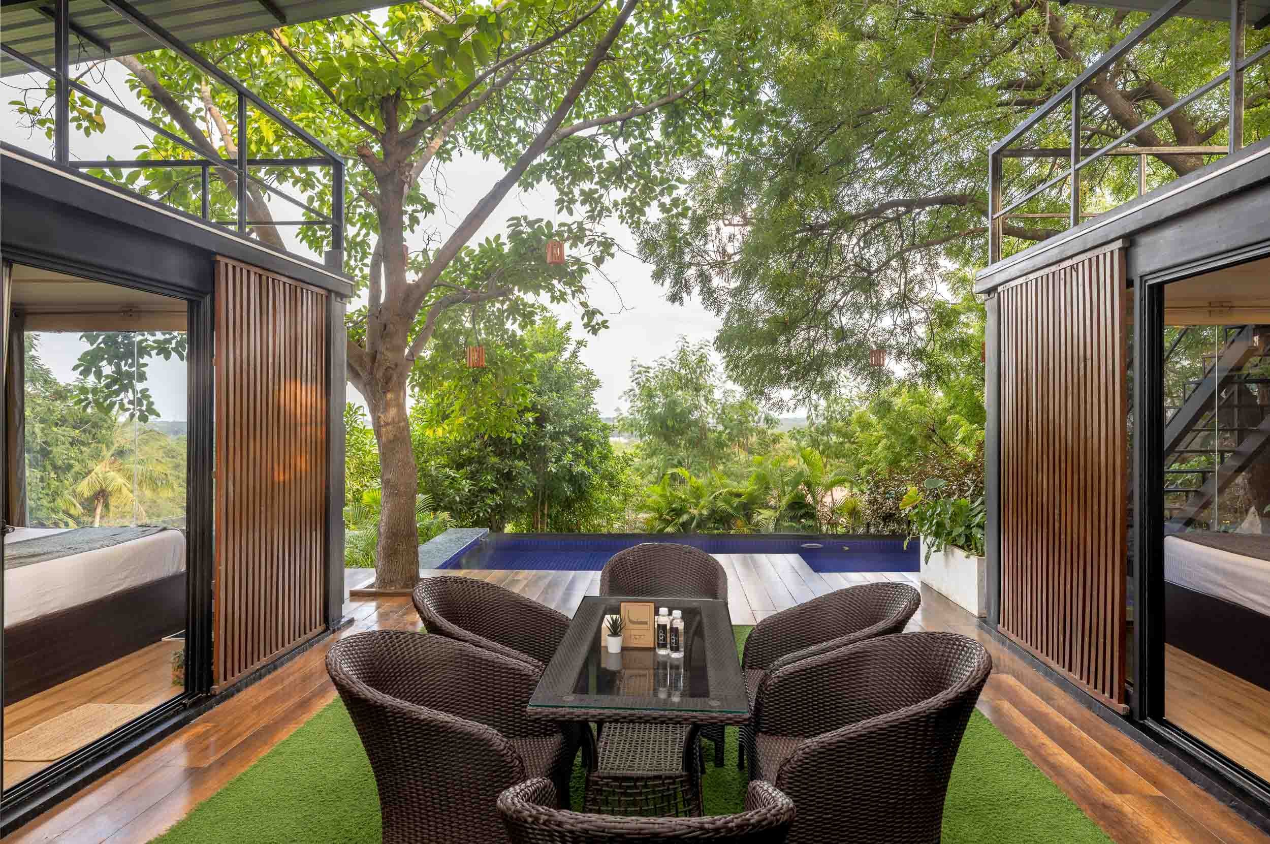 Exterior photography of a modern luxury staycation in Hyderabad, featuring a wooden deck with a private plunge pool, outdoor wicker seating, and lush green trees