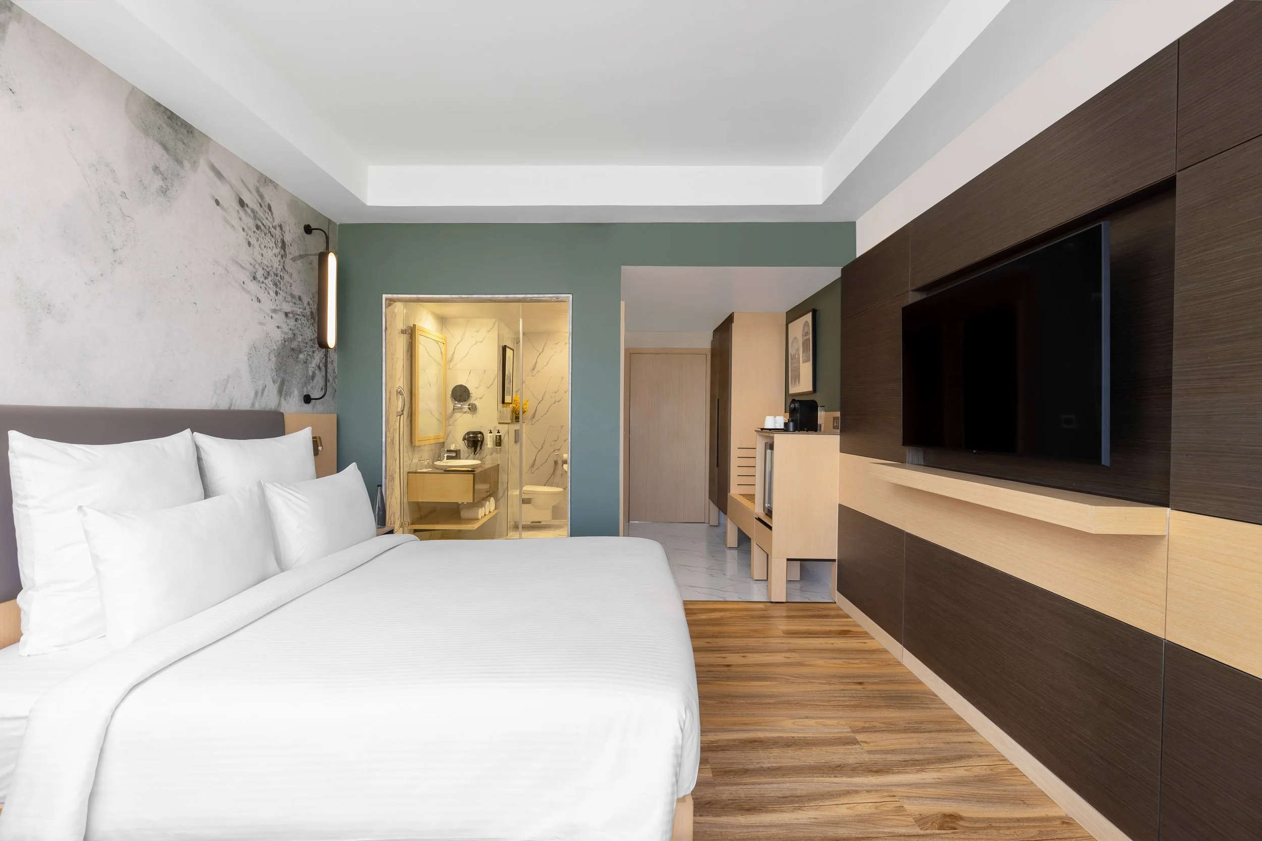 Interior photography of a modern hotel bedroom featuring a large bed, wooden flooring, a wall-mounted TV on a wooden panel, and a long desk console