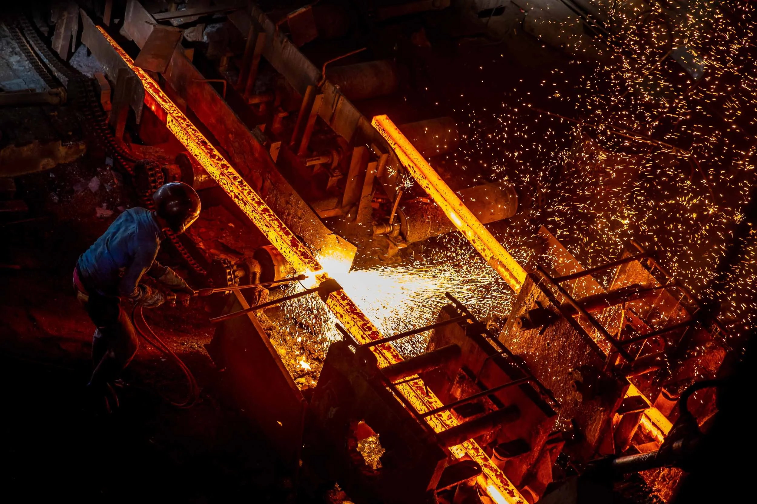 Dynamic industrial photography of a worker handling glowing molten metal amidst flying sparks in a heavy manufacturing foundry