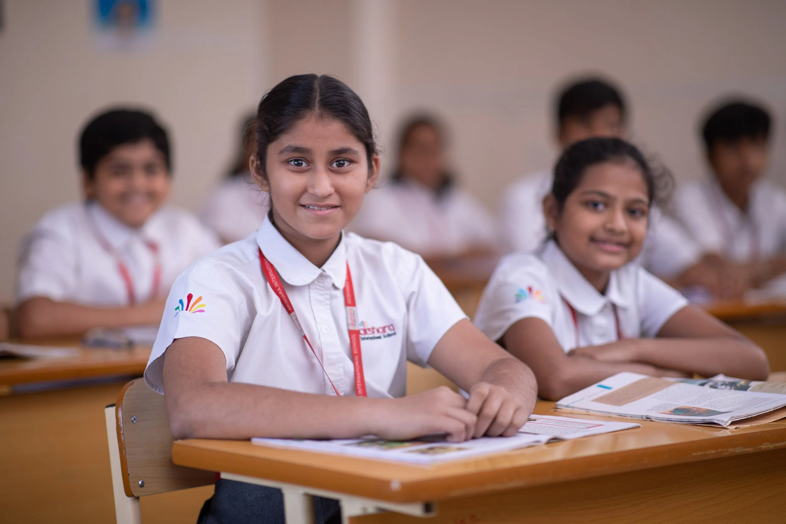 A group of young female students wearing white school uniforms with red lanyards, sitting together at wooden desks in a brightly lit classroom and smiling at the camera