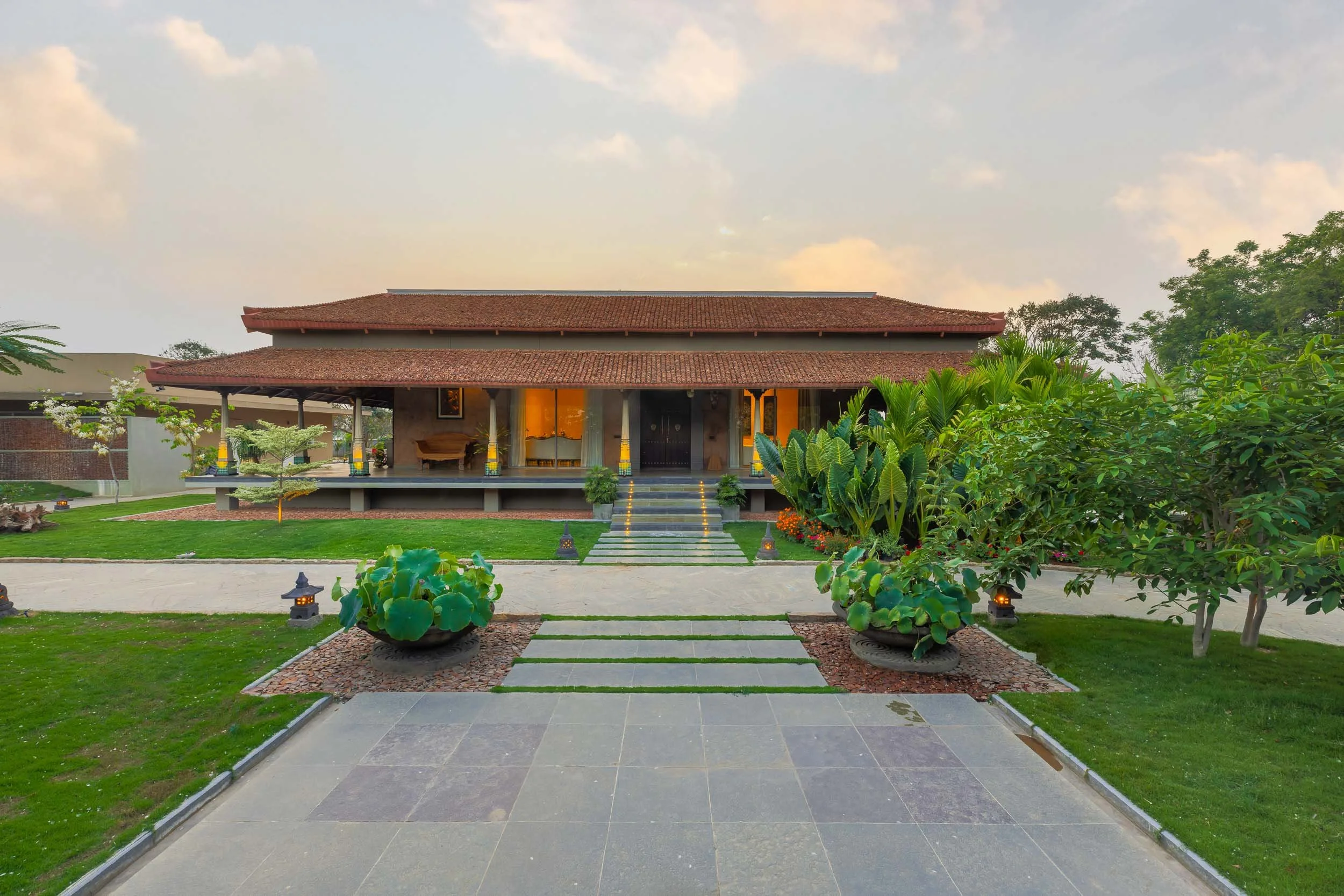 Wide exterior daytime photography of a luxury resort farm stay featuring a continuous covered veranda, a traditional pitched roof, and an expansive green lawn in the foreground