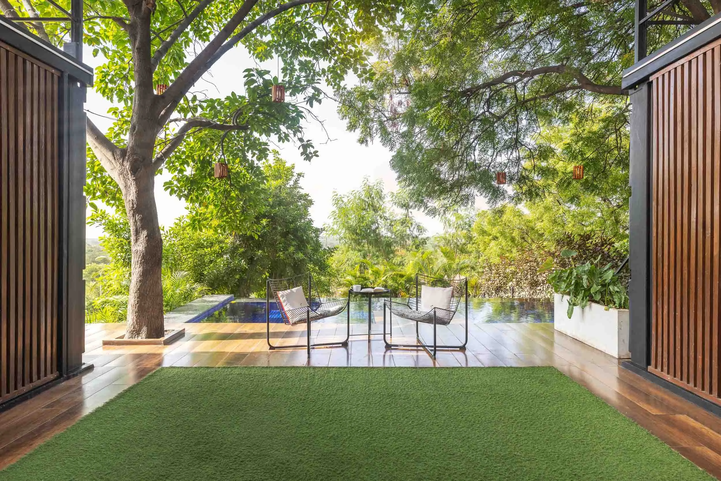 Exterior daytime photography of a modern luxury staycation in Hyderabad, featuring an elevated deck with glass railings, outdoor seating, and views of a lush green lawn