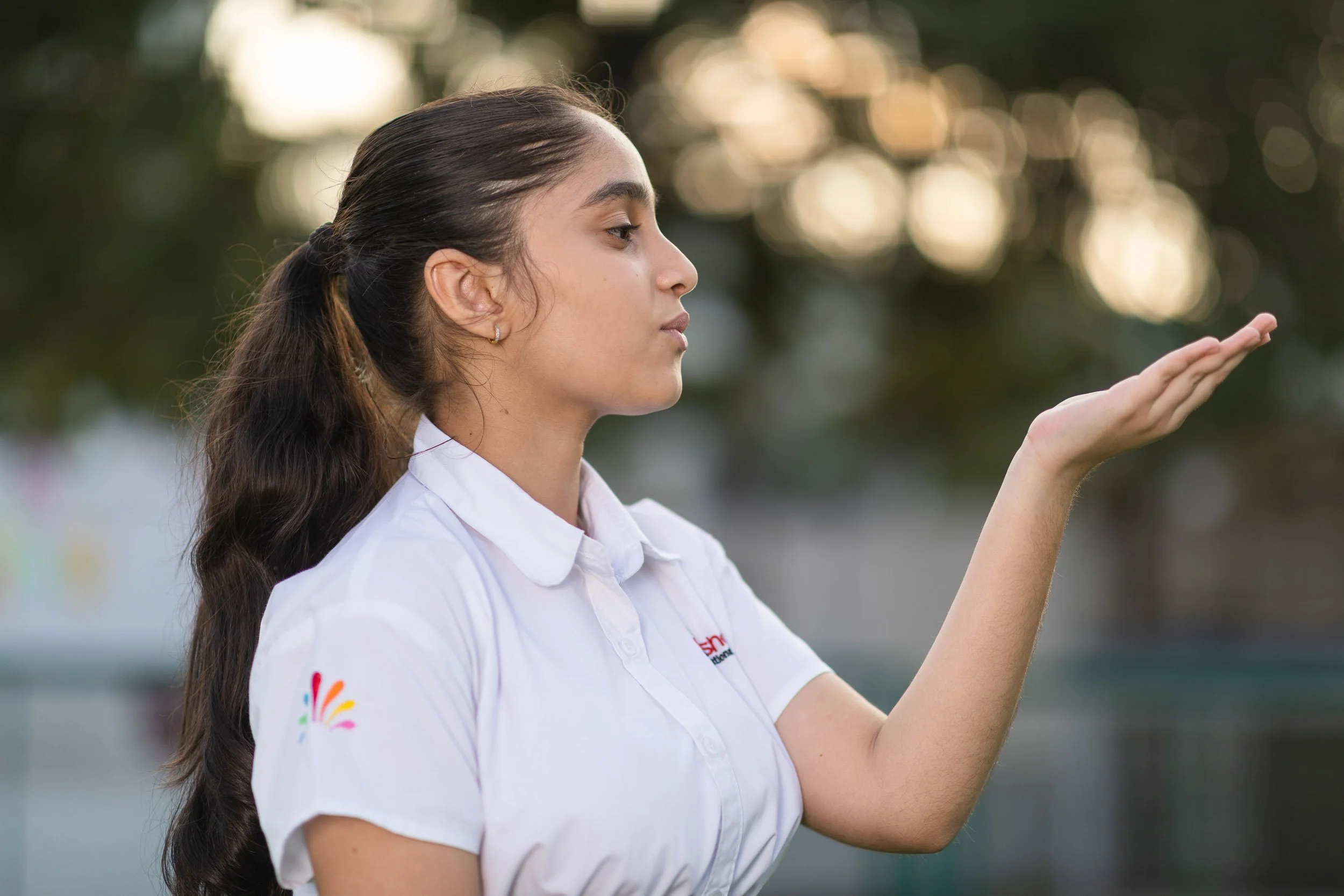 A young female student wearing a white uniform shirt with her hair in a ponytail, standing outdoors with a blurred green background. She is playfully holding her hand open near her face