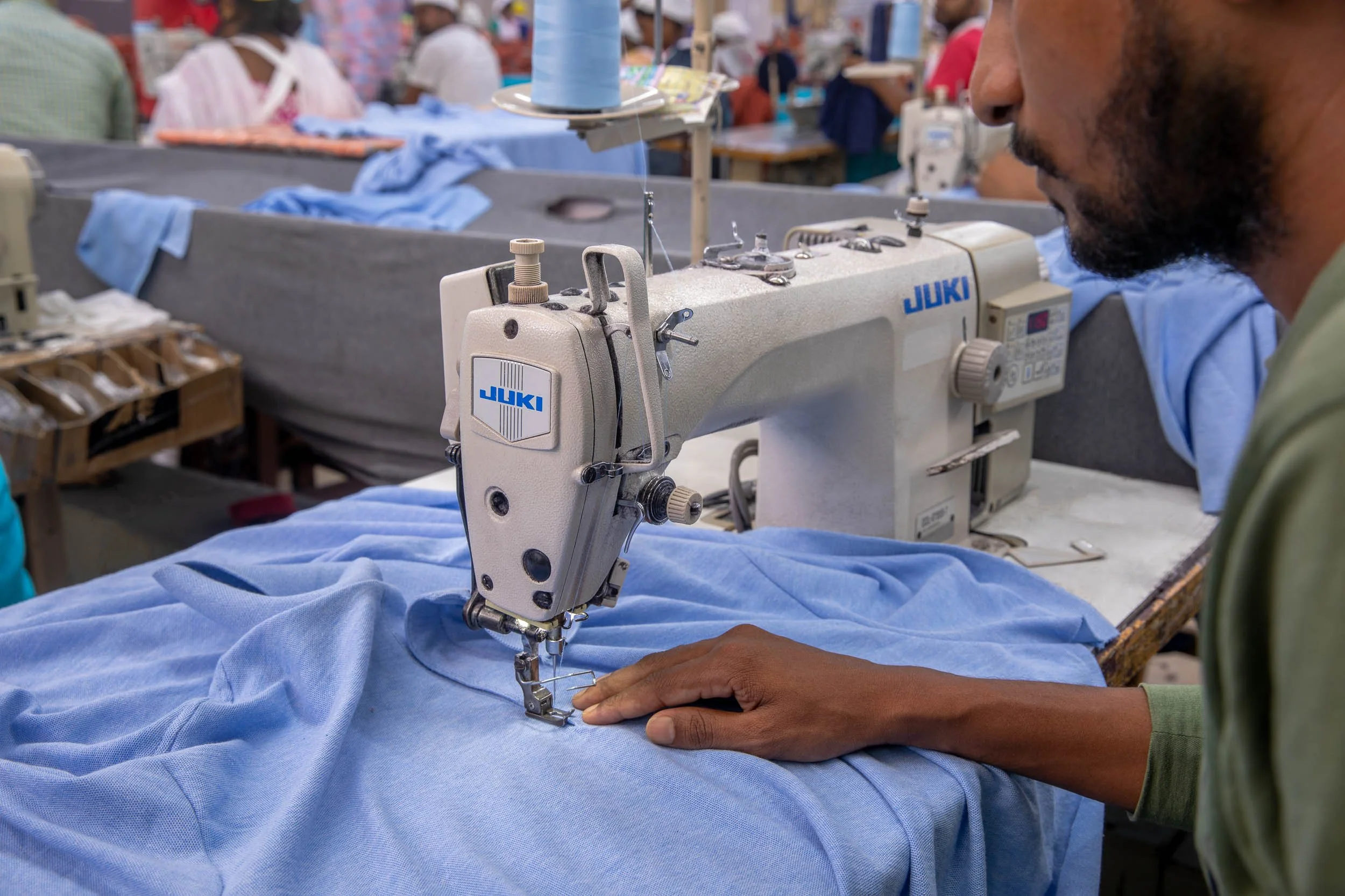 Close-up industrial photography of a garment worker operating a sewing machine on light blue fabric in a textile manufacturing plant