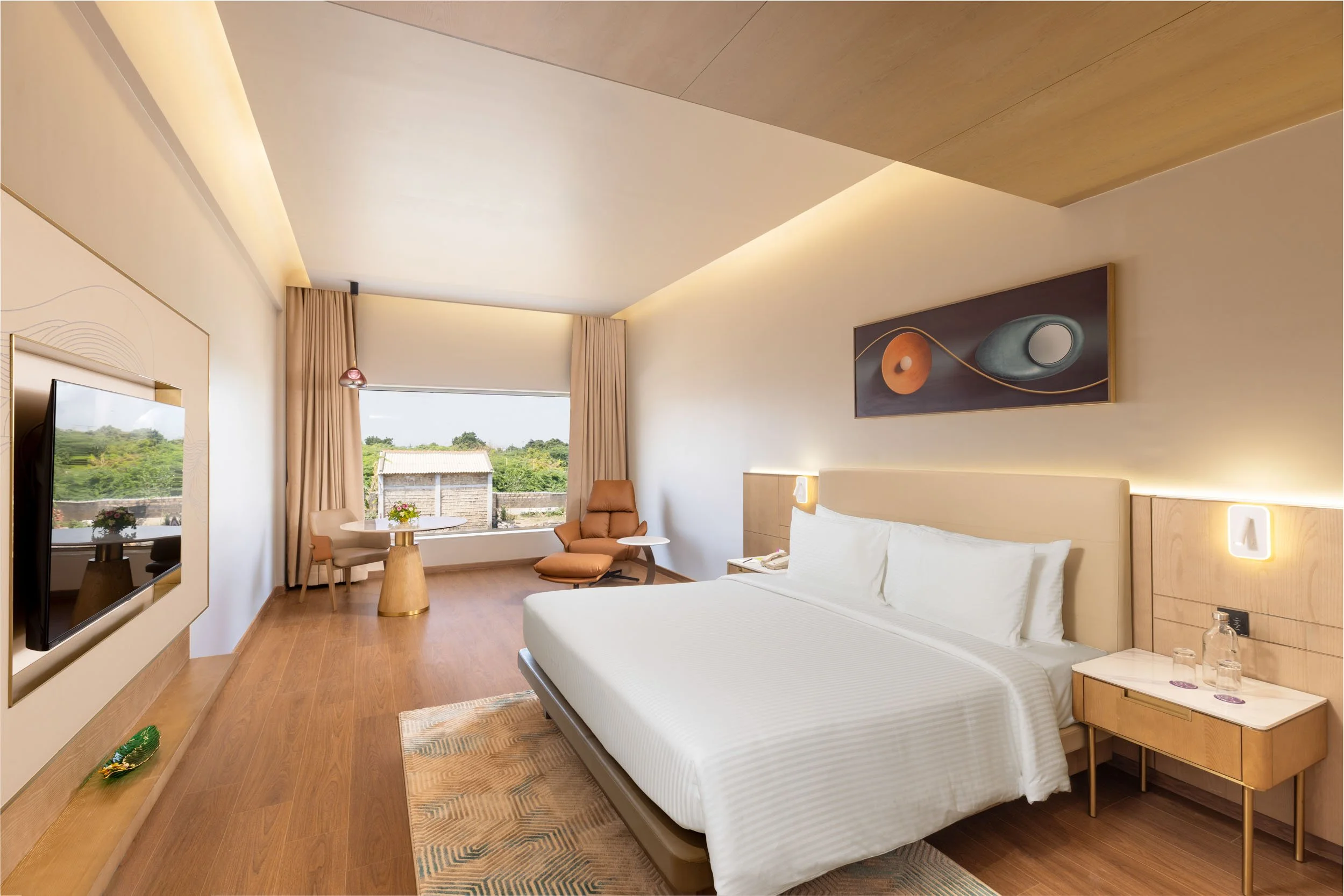 Interior photography of a spacious modern hotel room featuring a king bed, a long wooden desk console, a flat-screen TV, and a tray ceiling with warm lighting