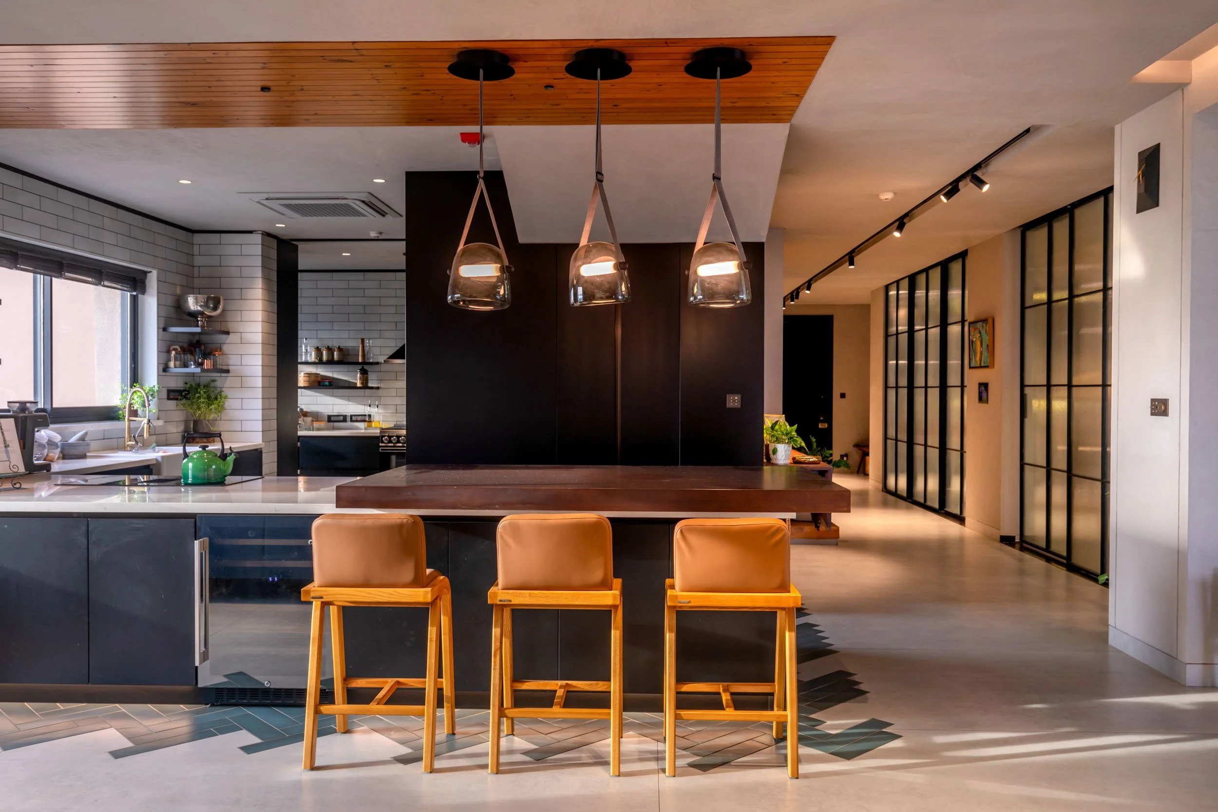 Modern open-plan kitchen and dining area featuring a dark island with orange leather stools, pendant lighting, and a wooden ceiling accent