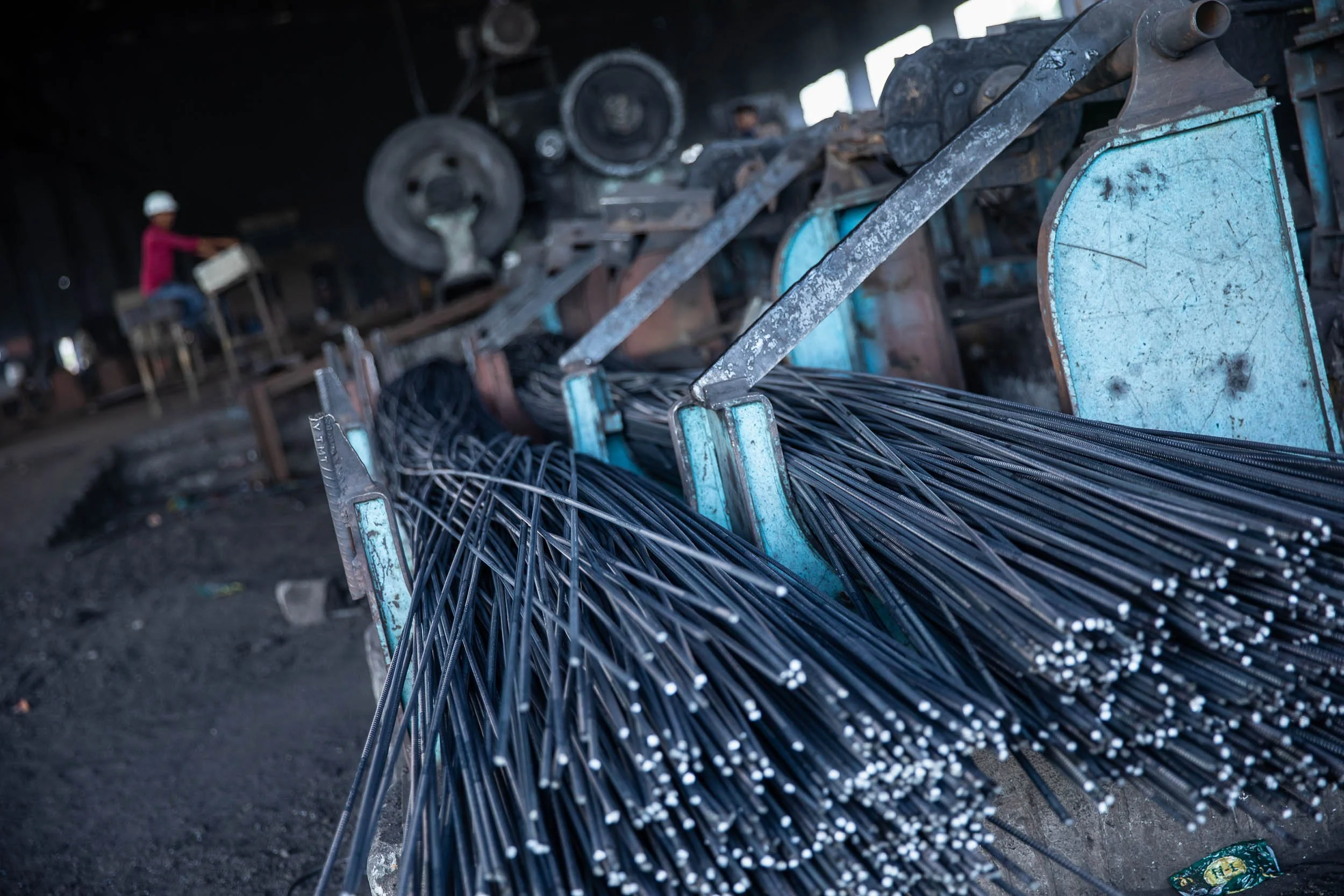 Interior industrial photography of a large stockpile of steel rebar on the floor of a heavy metal manufacturing plant, with a factory worker in the background.