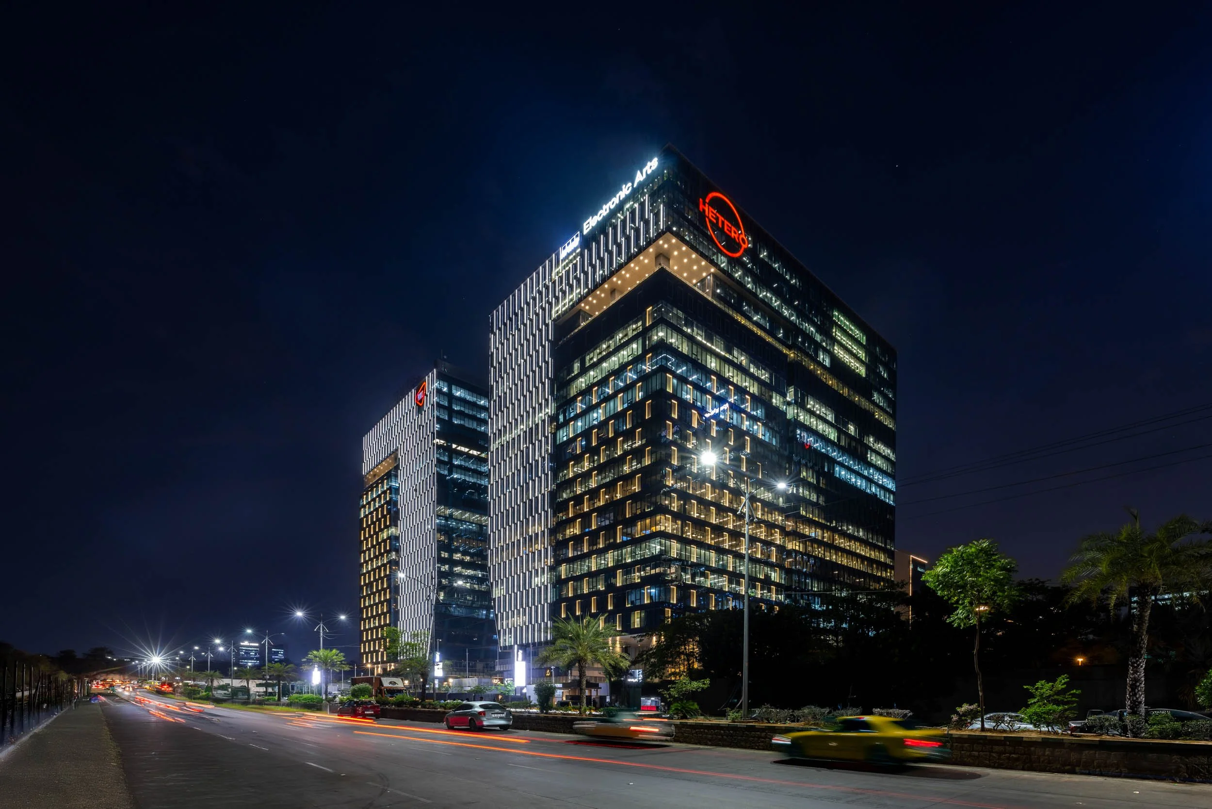 Professional commercial architecture photography capturing a striking modern office building at twilight, highlighting the glowing glass facade and dynamic urban energy