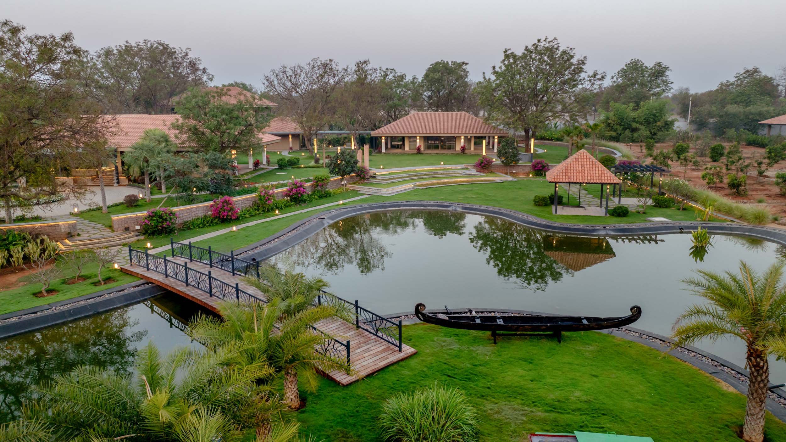 Resort landscape photography featuring a tranquil pond with a traditional wooden boat, surrounded by lush green lawns and tiled-roof villas