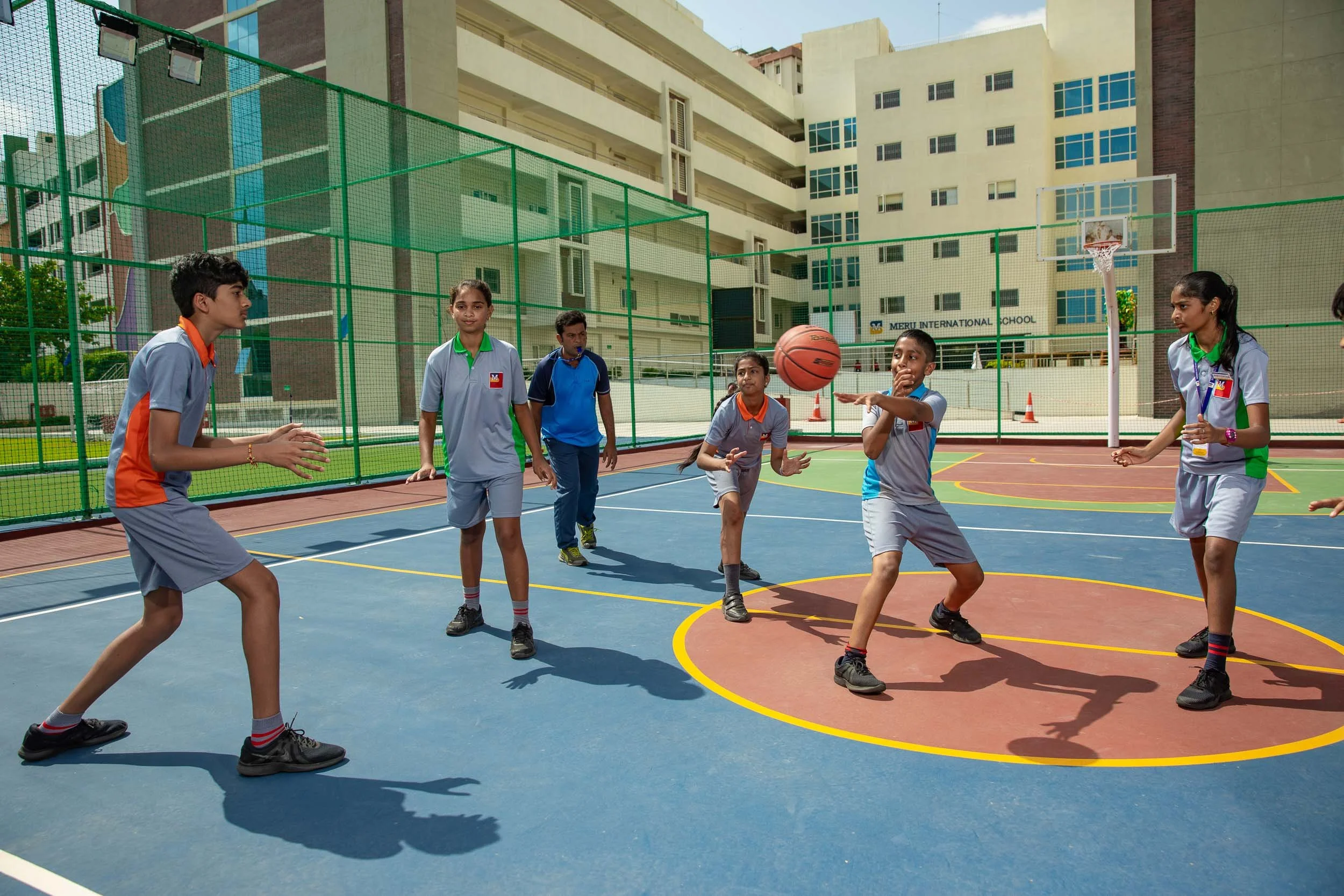 A group of students in school uniforms actively playing a game of basketball on a bright blue and red outdoor court, with a large, multi-story school building in the background