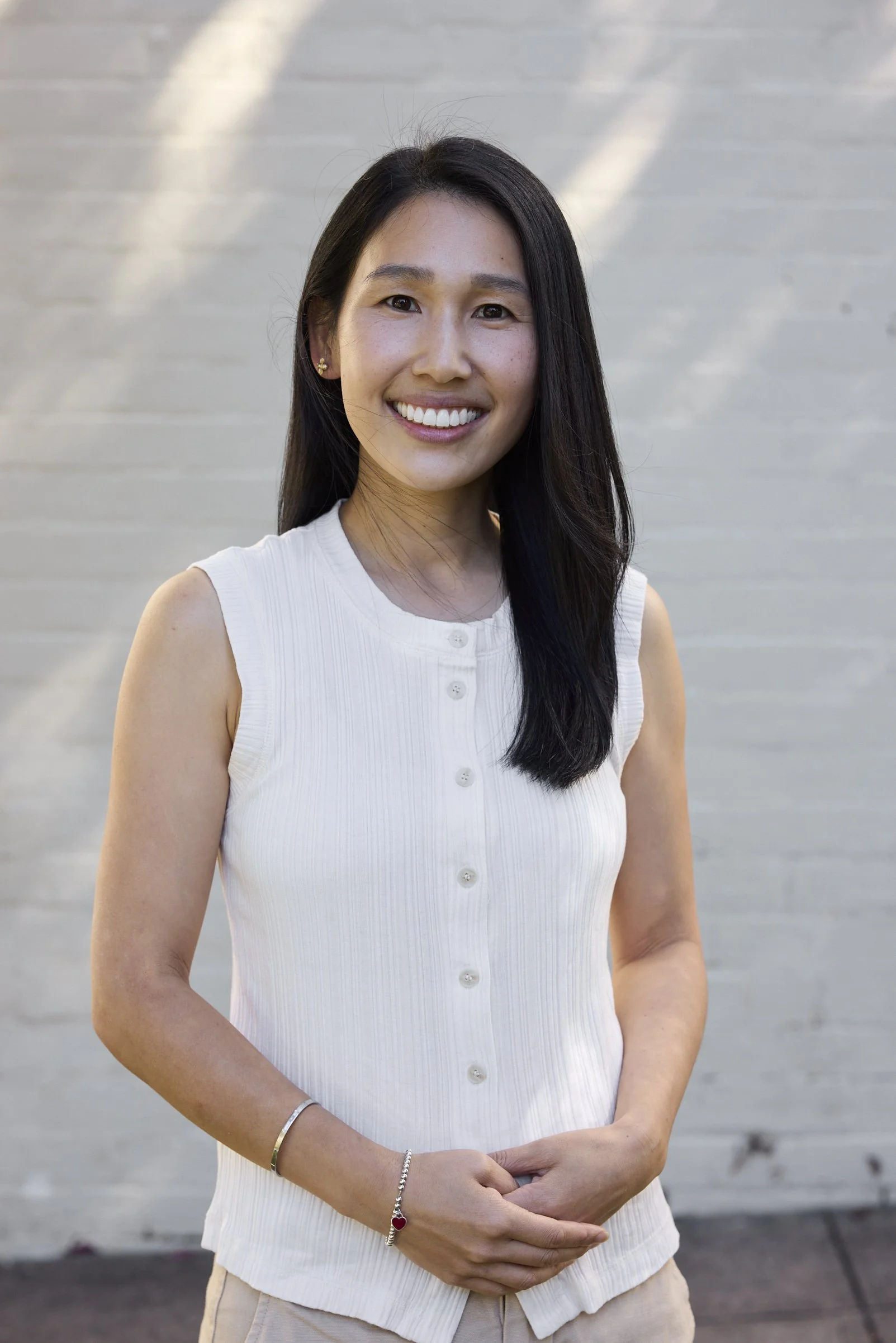 A smiling woman with long black hair wearing a sleeveless white button-up top, standing outdoors against a white brick wall.