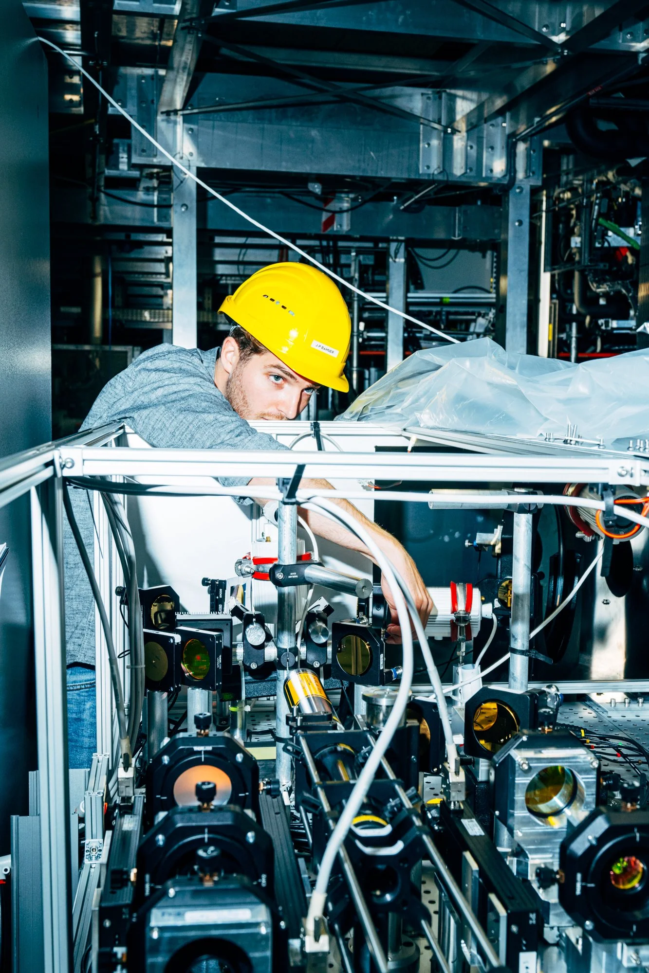 Ein Mann trägt einen gelben Schutzhelm und arbeitet an einem komplexen wissenschaftlichen oder technischen Gerät mit verschiedenen mechanischen und elektronischen Komponenten in einem Labor.
