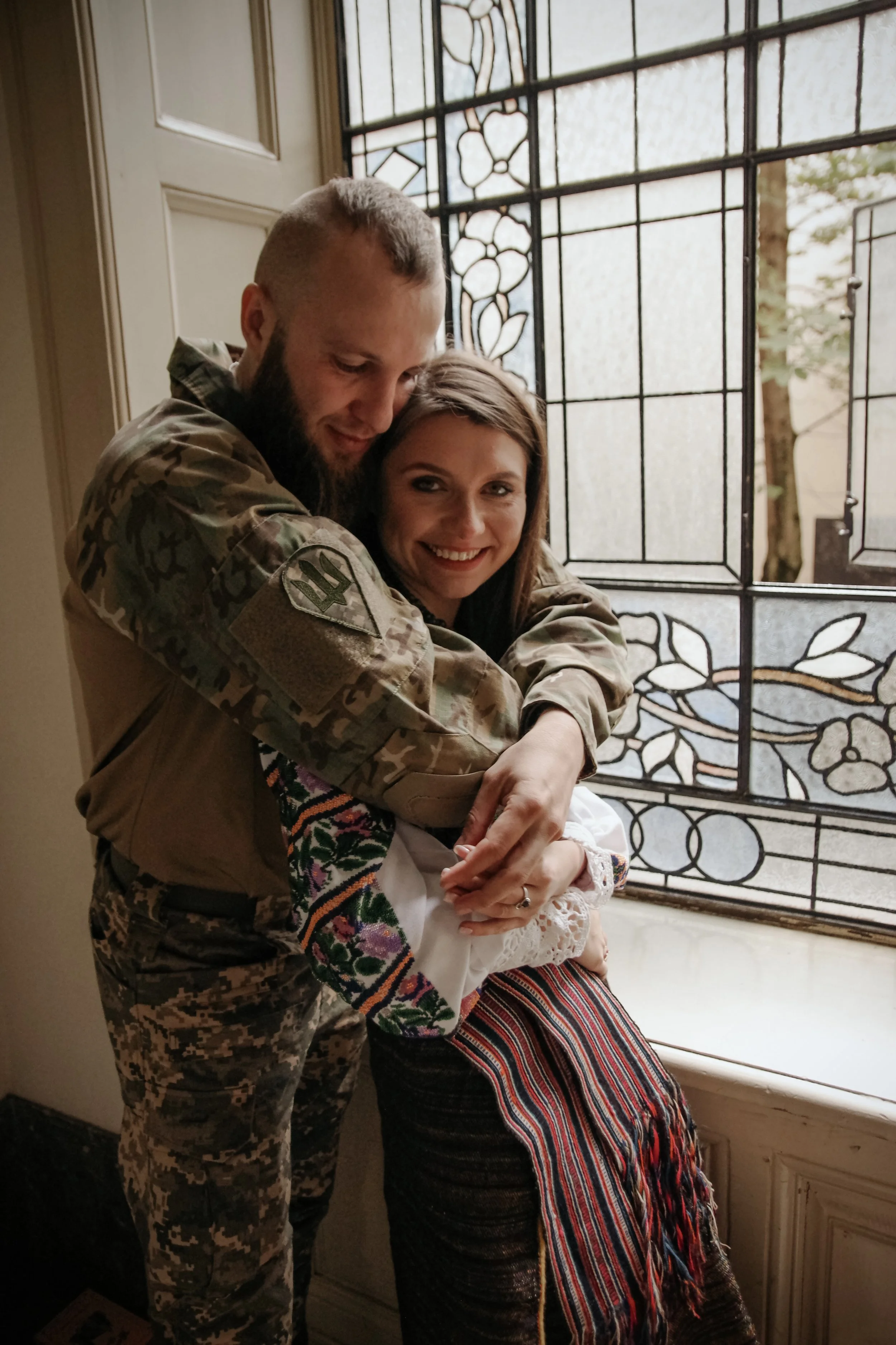 Ein Mann in Camouflage-Uniform umarmt und hält ein lächelndes Mädchen in einem bunten, traditionell bestickten Kleid vor einem kunstvoll verzierten Fenster.