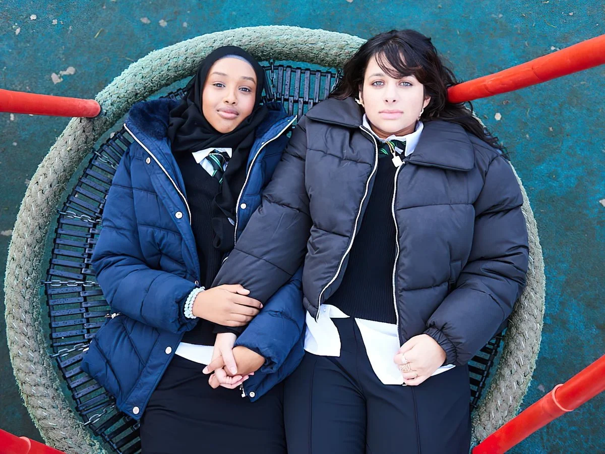 Two girls lying on a round netted platform at a playground, holding hands. One girl is wearing a hijab and a blue jacket, and the other girl has dark hair and is wearing a black jacket.