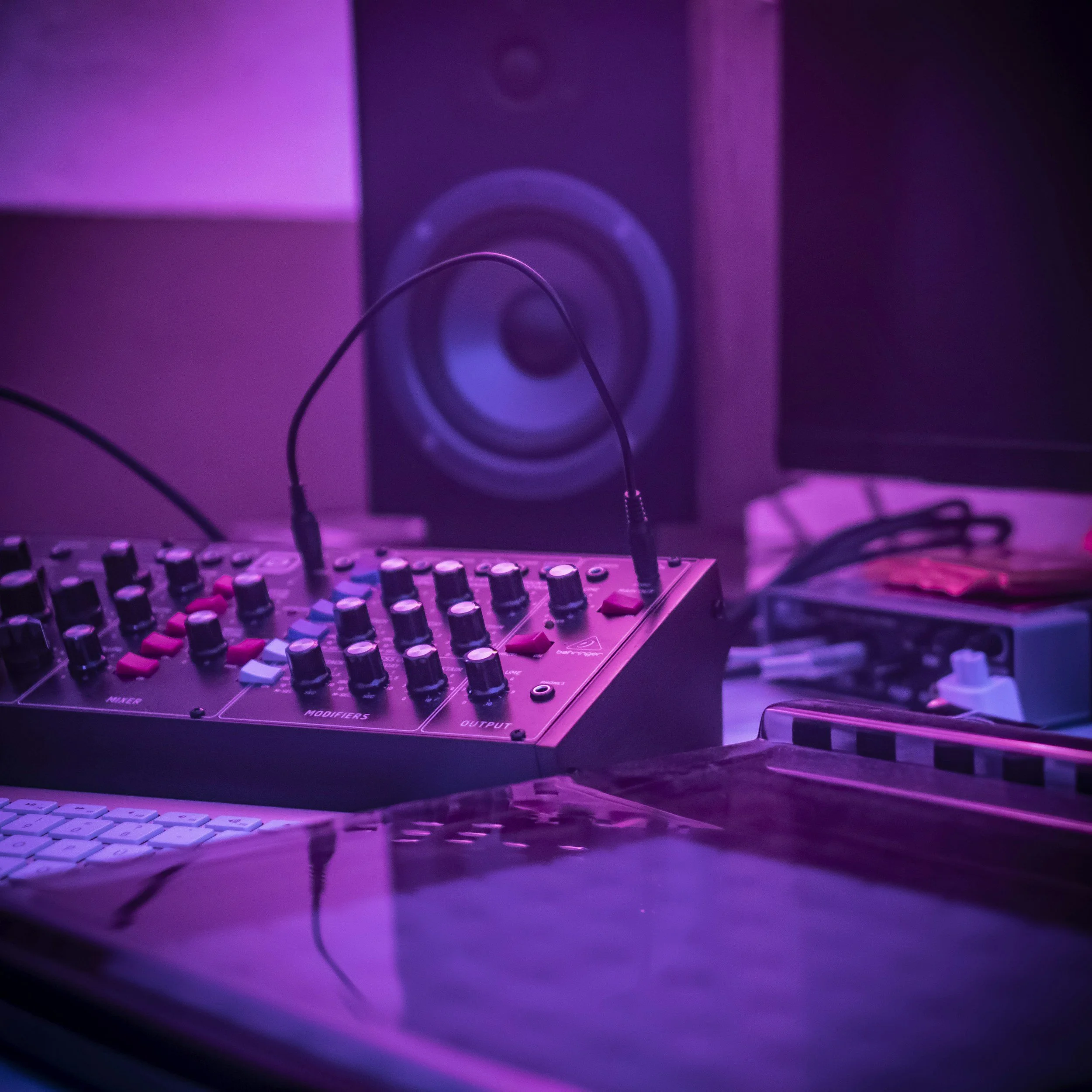 Close-up of a DJ equipment setup in a room illuminated with purple and pink lighting, featuring knobs, sliders, and cables connected to audio equipment.