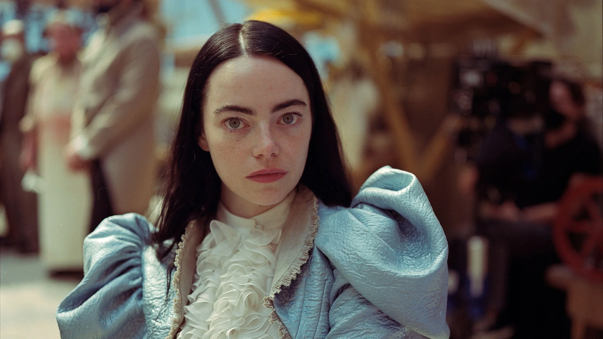 A woman with pale skin and black hair, wearing a blue dress with a ruffled white collar, looking directly at the camera in a store or boutique with mannequins and clothing displays in the background.