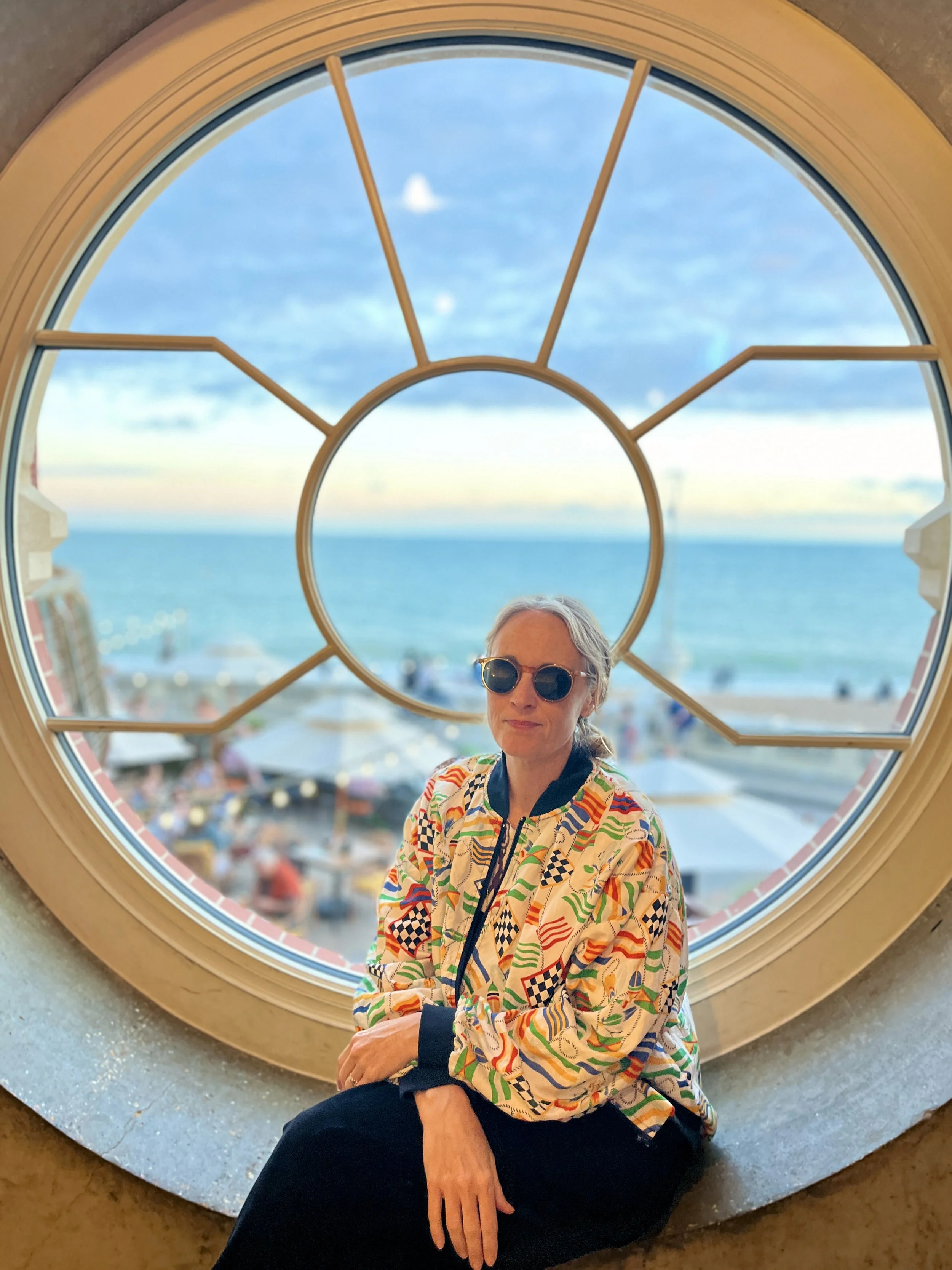 A woman with gray hair and sunglasses sitting on a circular window sill with a view of the ocean and sky in the background.