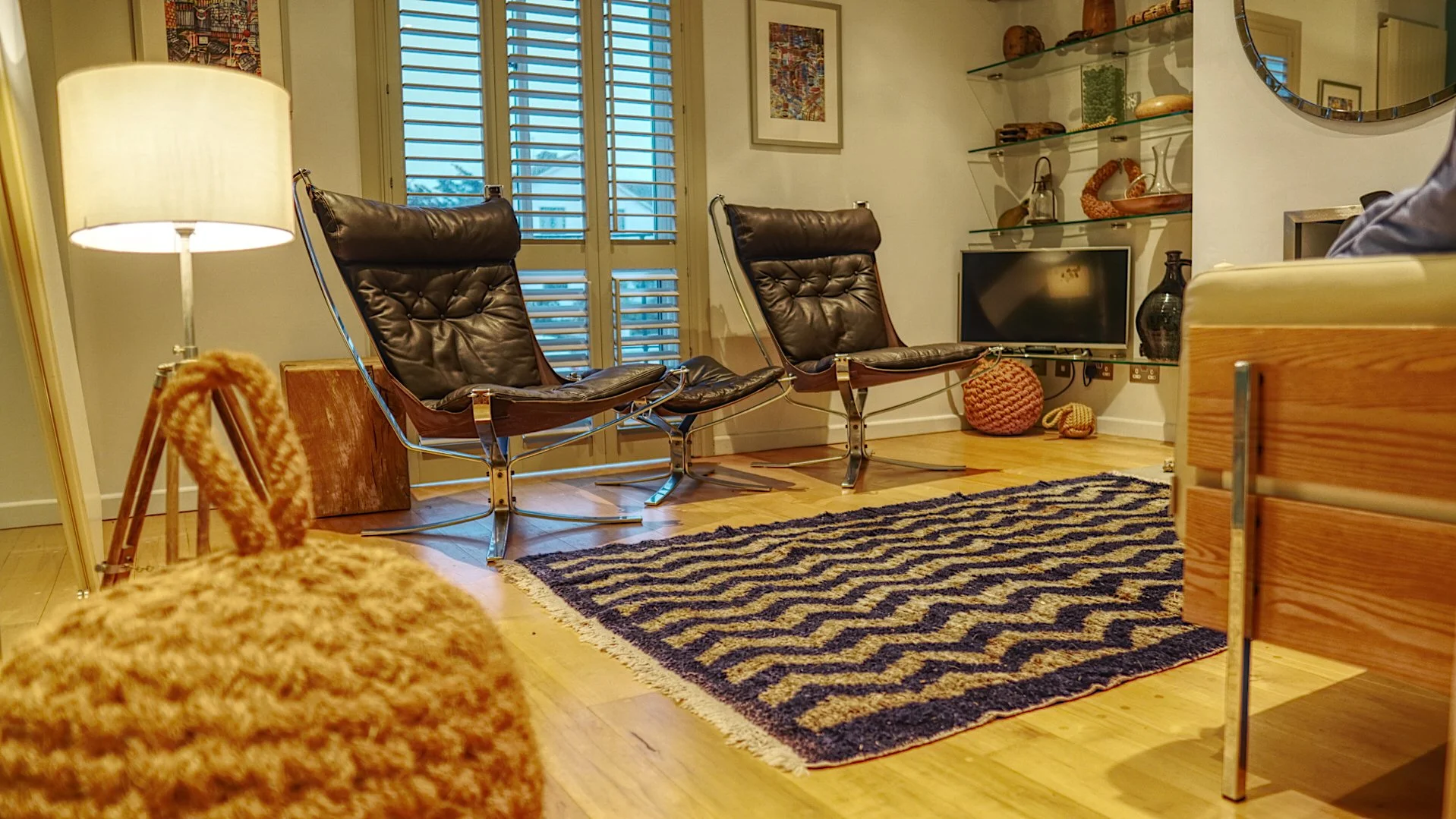 Living room with two modern brown leather chairs, a black and white striped rug, a flat-screen TV, and shelves with decorative items, plaid curtains on windows, and framed artwork on the walls.