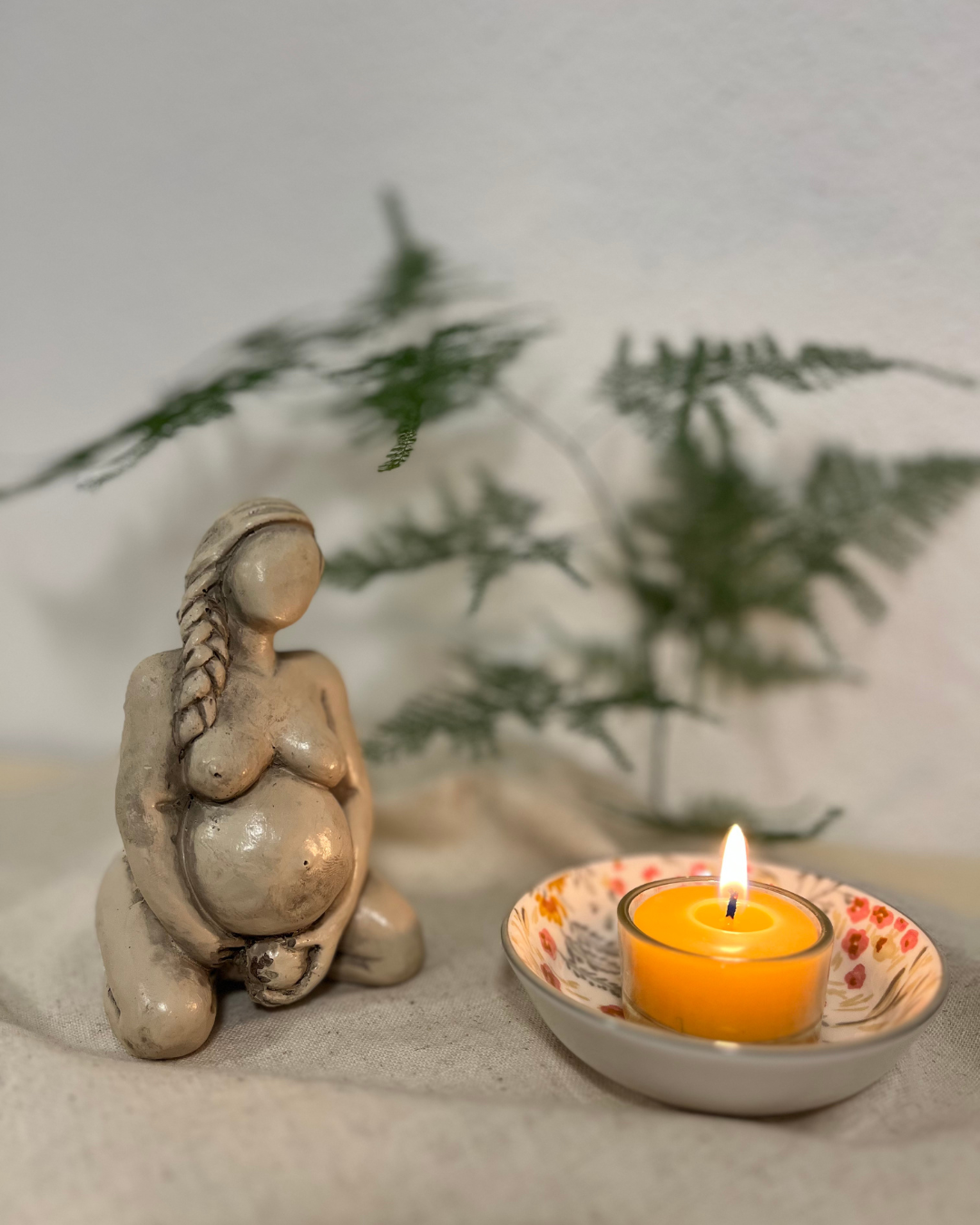 Eine ceramic Figur einer schwangeren Frau mit langer, geflochtener Frisur, neben einer Kerze in einem dekorativen Schälchen, im Hintergrund grüne Zweige auf weißem Stoff.