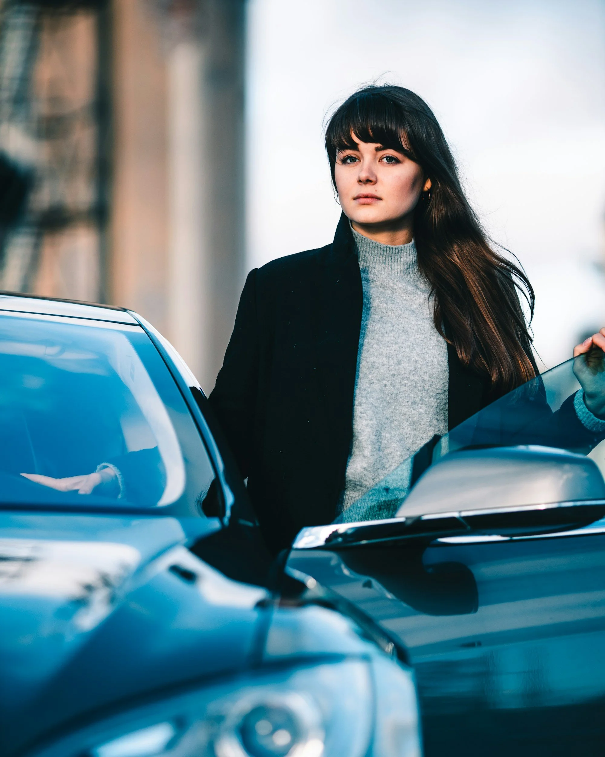 A woman with long dark hair and bangs standing beside a parked black car, wearing a light gray turtleneck and a black coat, looking confidently at the camera.