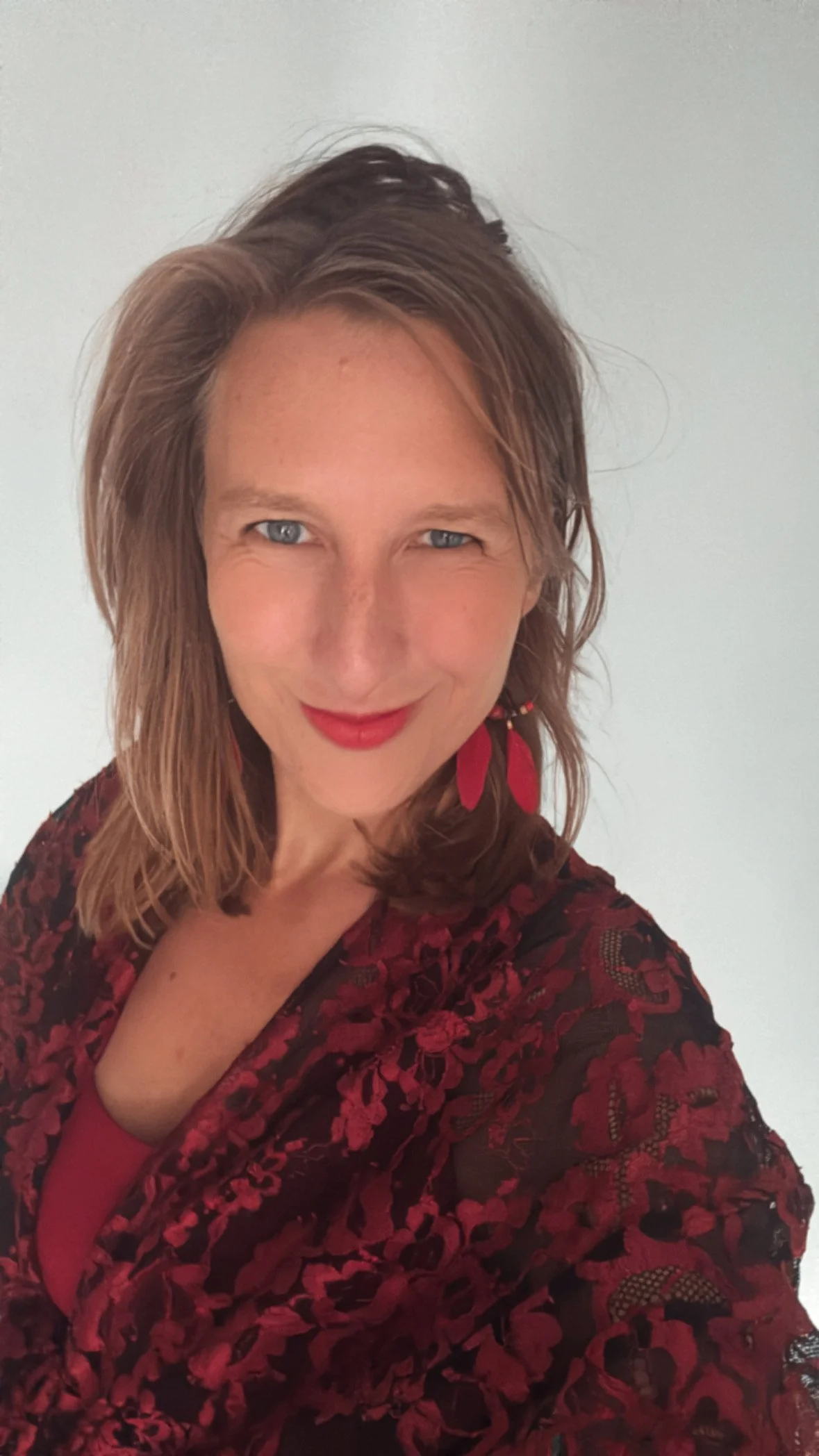 Woman with shoulder-length wavy brown hair, blue eyes, wearing red lipstick, red feather earrings, and a red and black lace patterned top, smiling against a plain white background.
