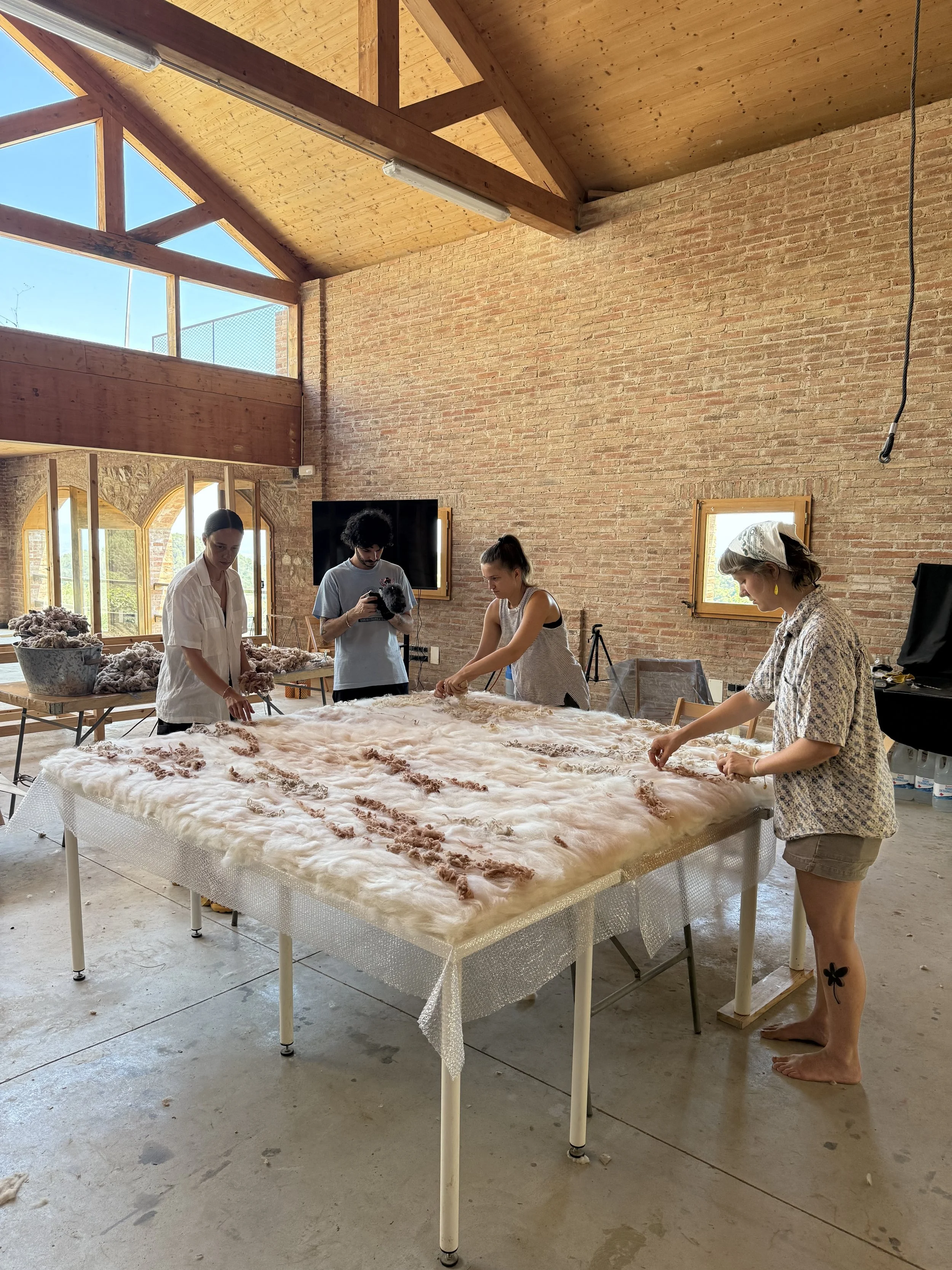 People working with wool on a large table in a bright room with brick walls and large windows.