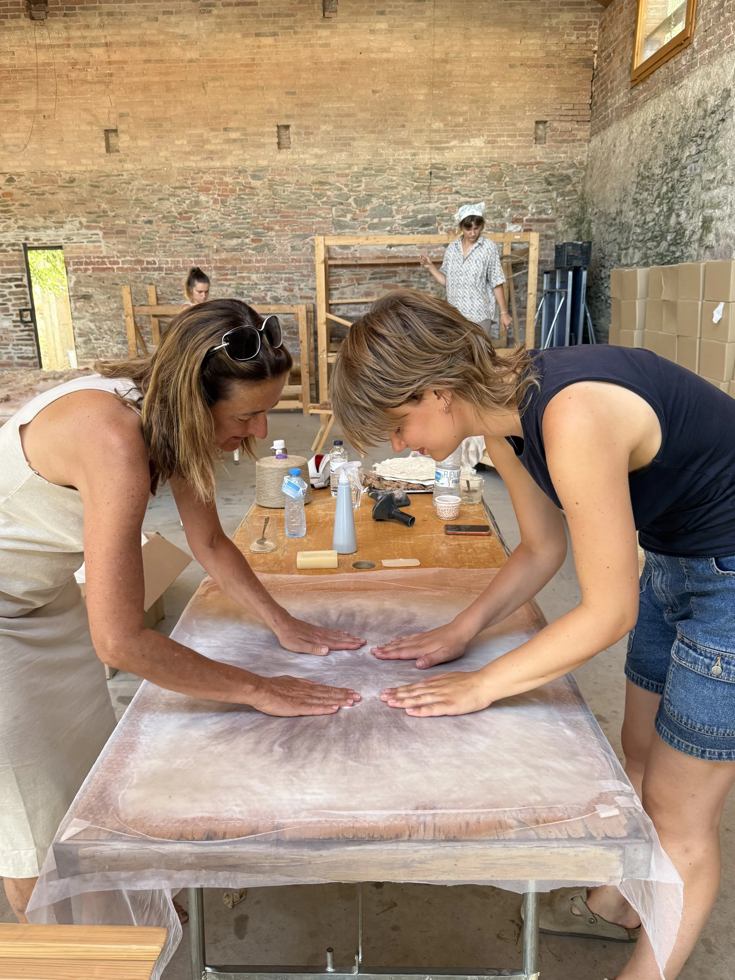 Two women bending over a table, working with dough, with spray bottles and kitchen supplies on the table. In the background, two women are working on a construction project in a room with brick walls and a window.