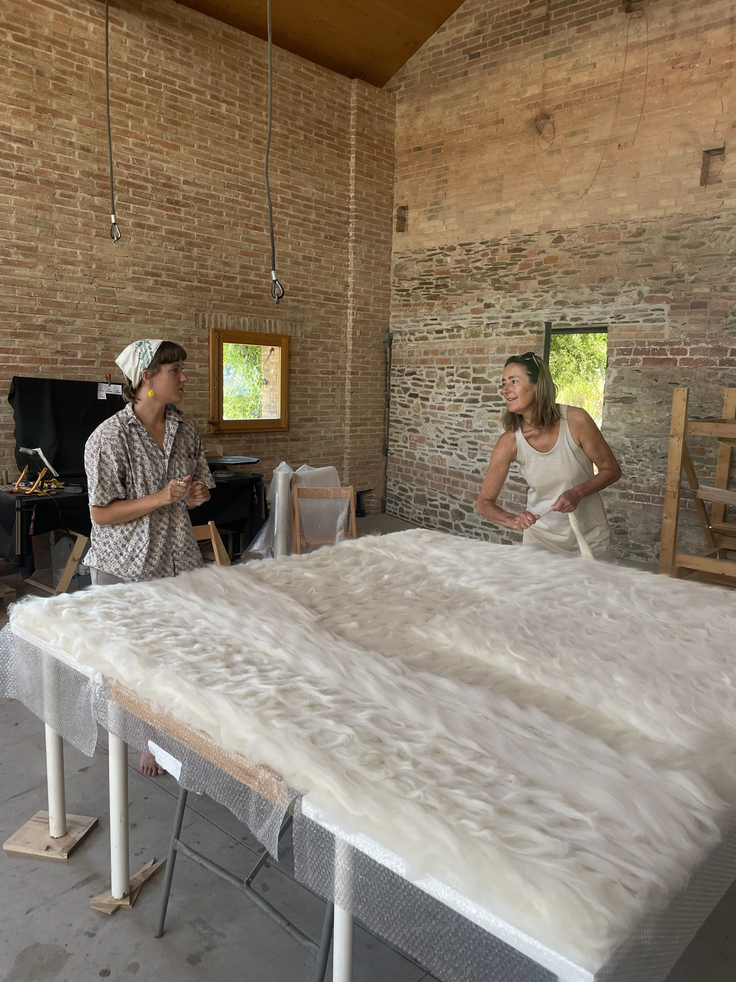 Two women with short hair and wearing casual clothing, one with a headscarf, standing near a large table covered with a white fluffy material inside a brick-walled room with small windows and hanging ropes.