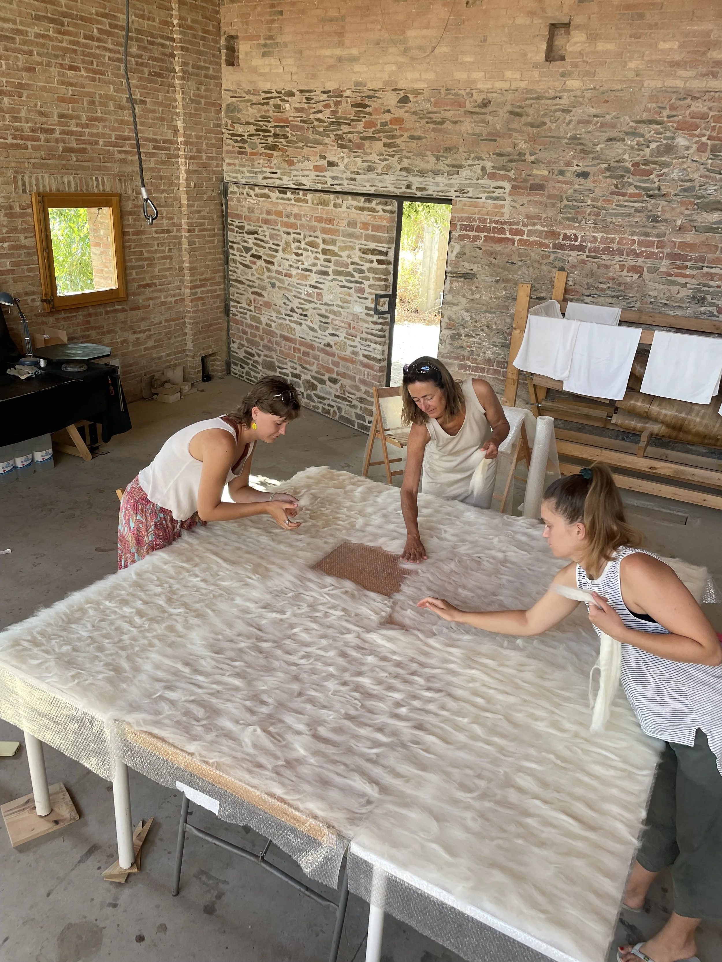 Four women are working on a large textured fabric draped over a table in an industrial space with exposed brick walls and a concrete floor. Three women are standing and leaning over the fabric, and one woman is reaching toward the fabric on the right