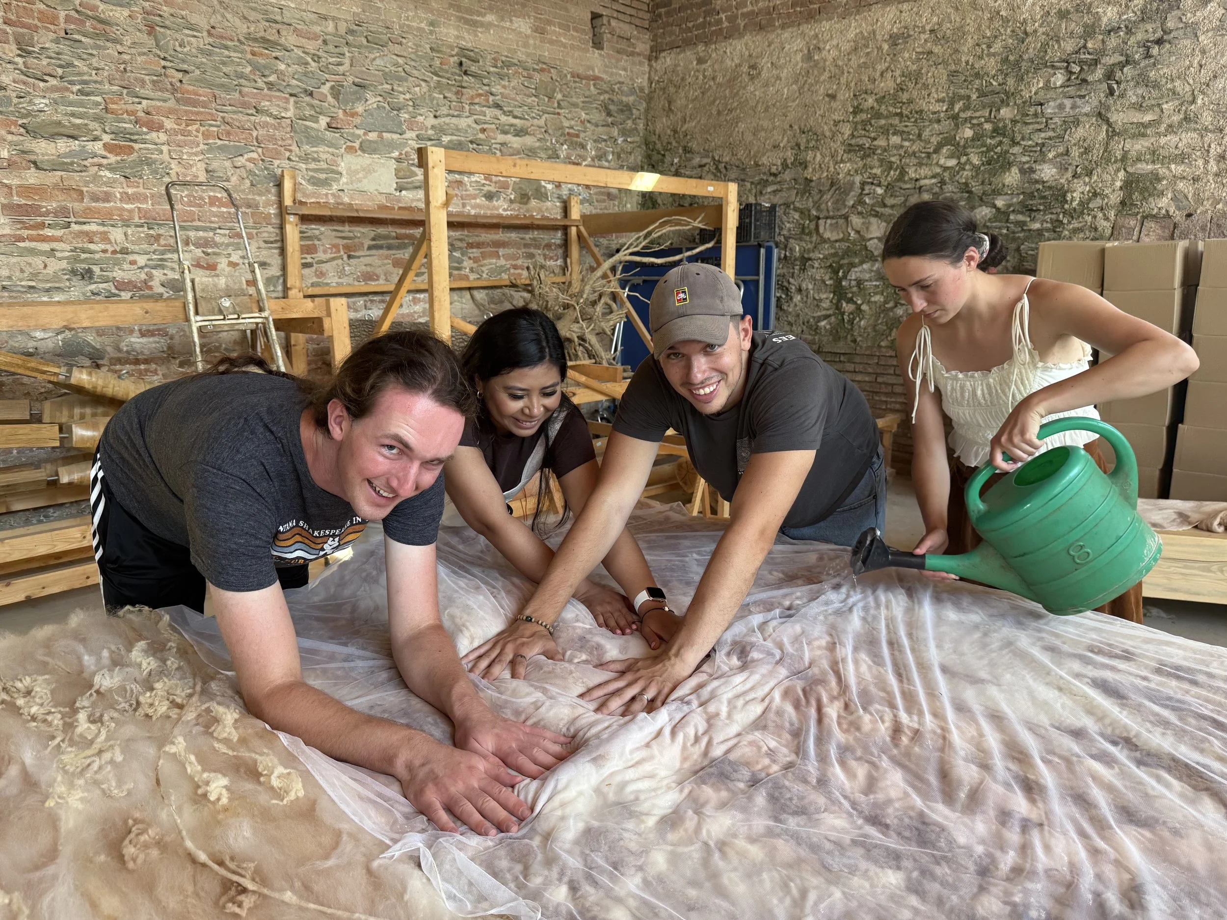 Group of five people kneeling on a large cloth in a brick-walled room, working together on a project, with one woman watering something with a green watering can, and various wooden structures and tools in the background.