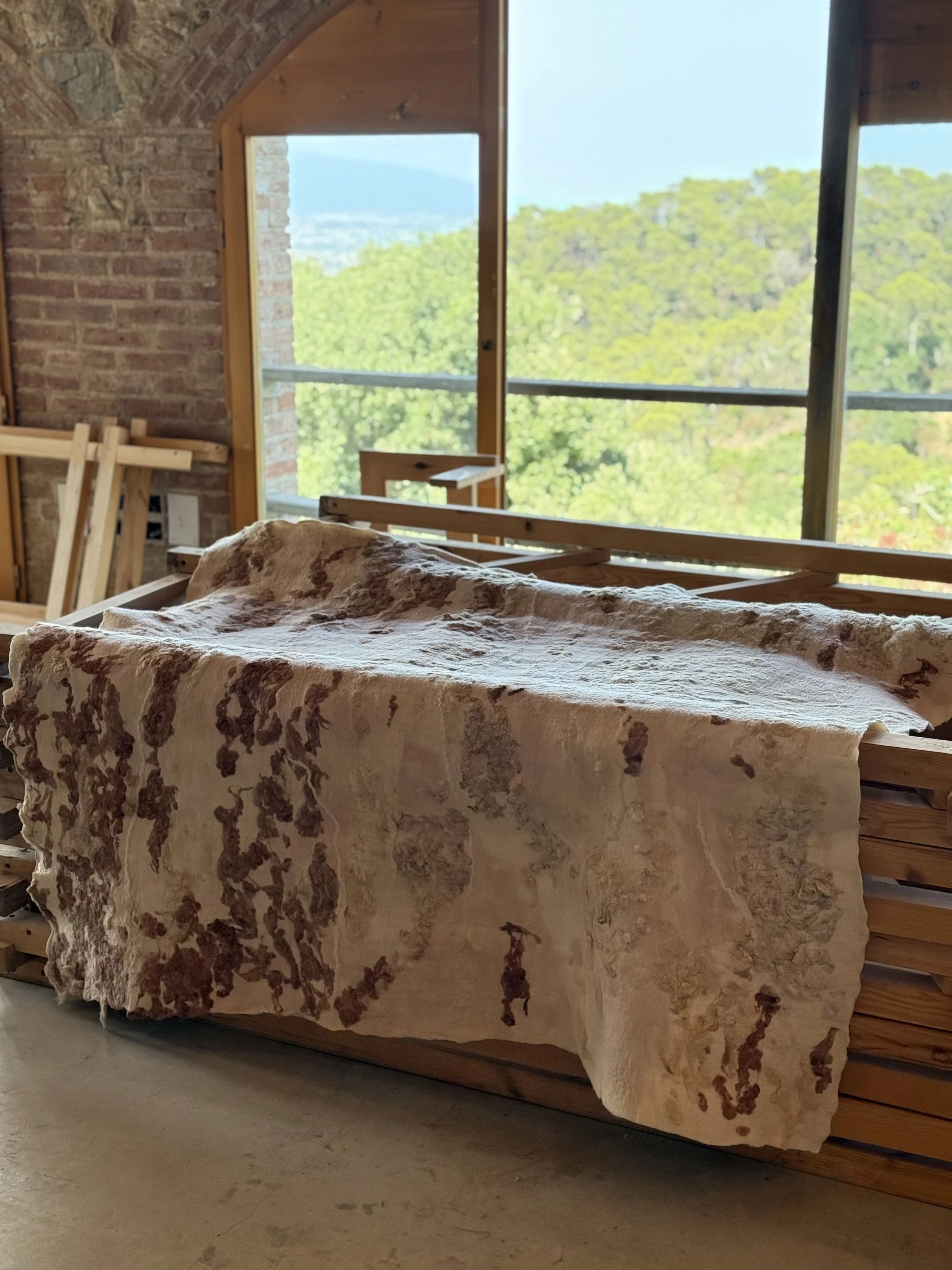 Unfinished wooden bed frame with a patterned fabric draped over it, set against a wall with a large window showing a view of trees and mountains outside, in a rustic room with brick and wood interior.
