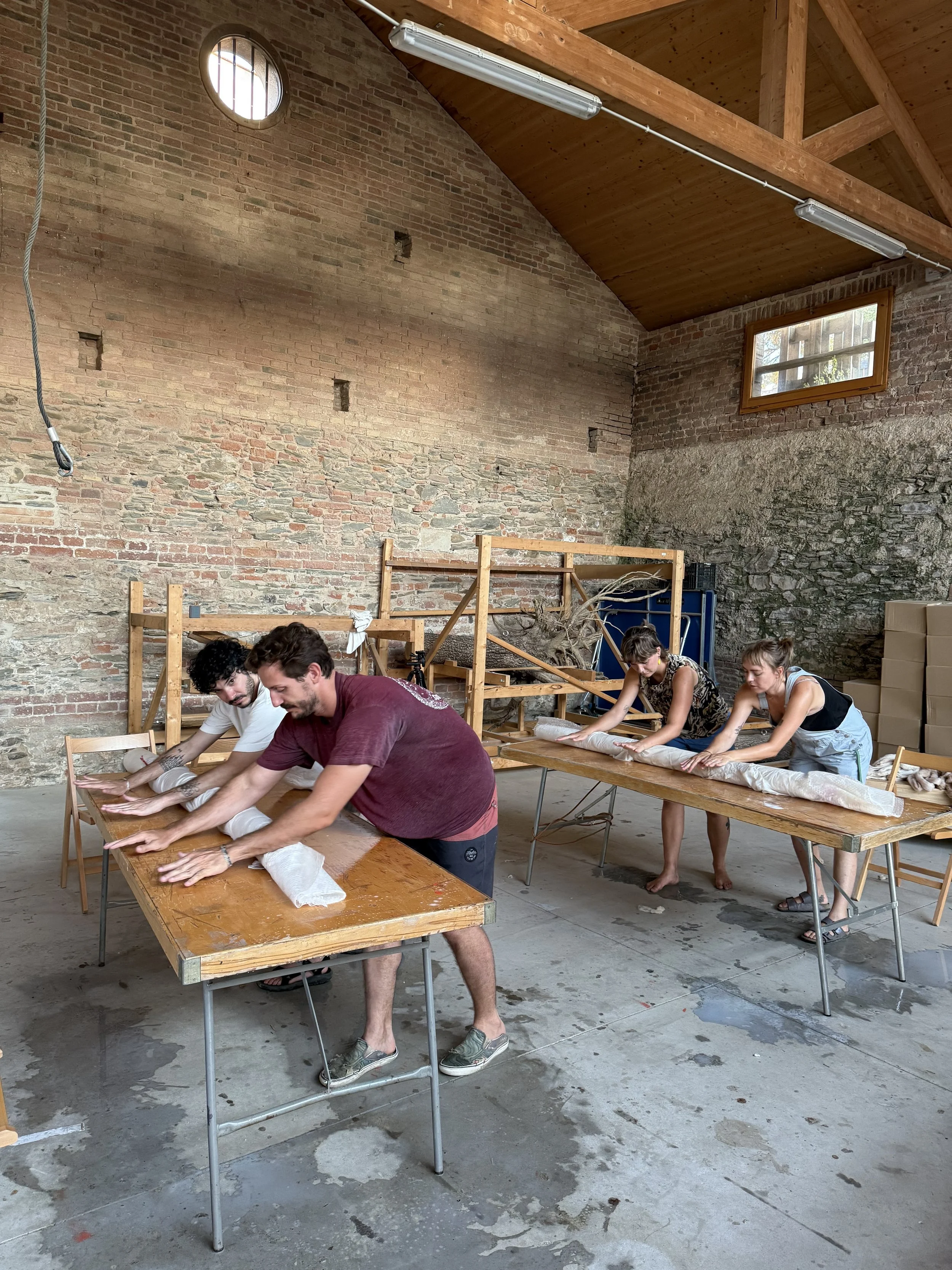 People seated at tables folding fabric or cloth in an industrial-style room with brick walls and wooden ceiling beams.