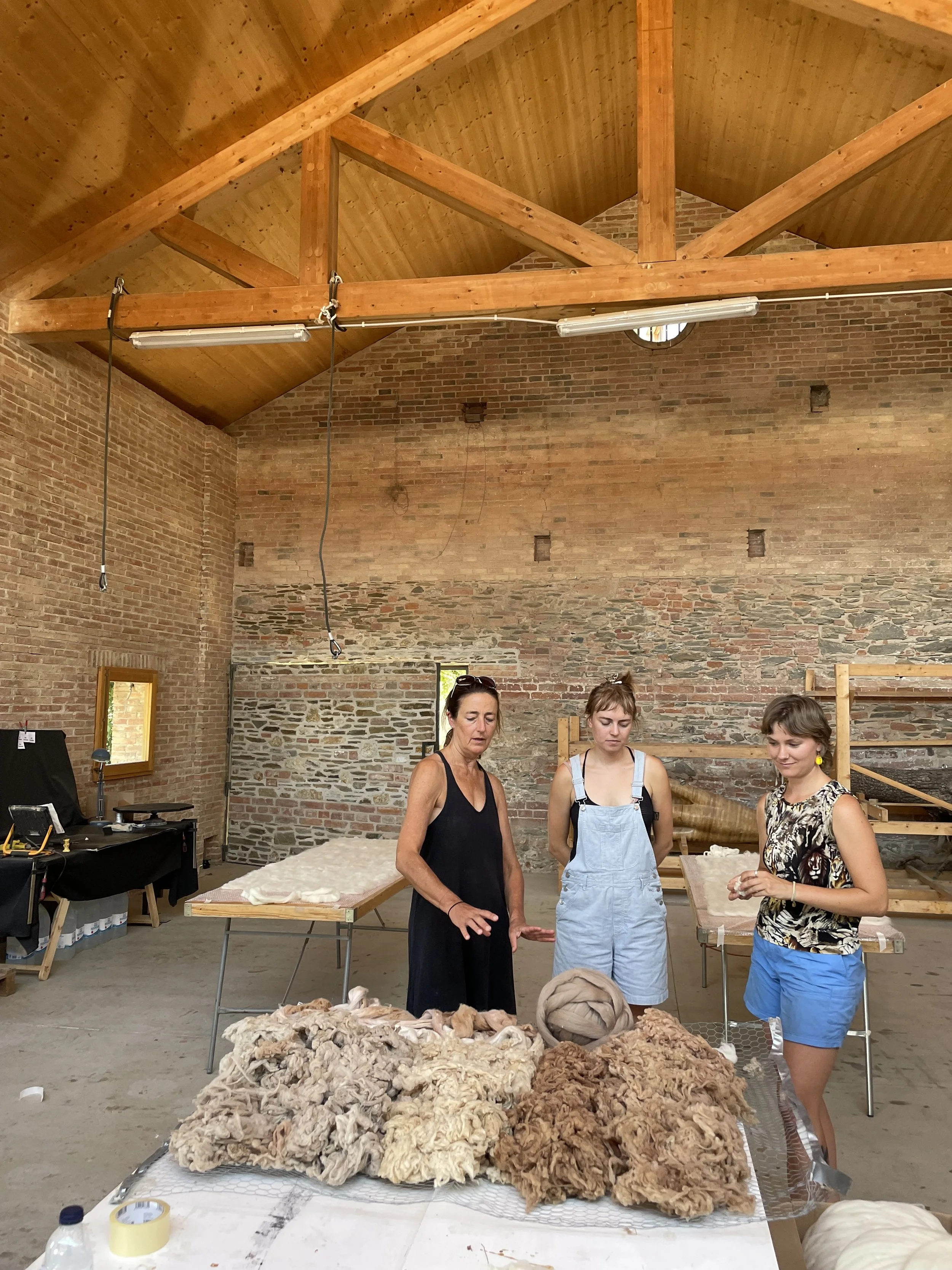 Three women standing behind a table with piles of raw wool or fleece in an indoor space with brick walls and wooden ceiling beams. Two of the women are looking down, and the third is holding a small object and smiling.