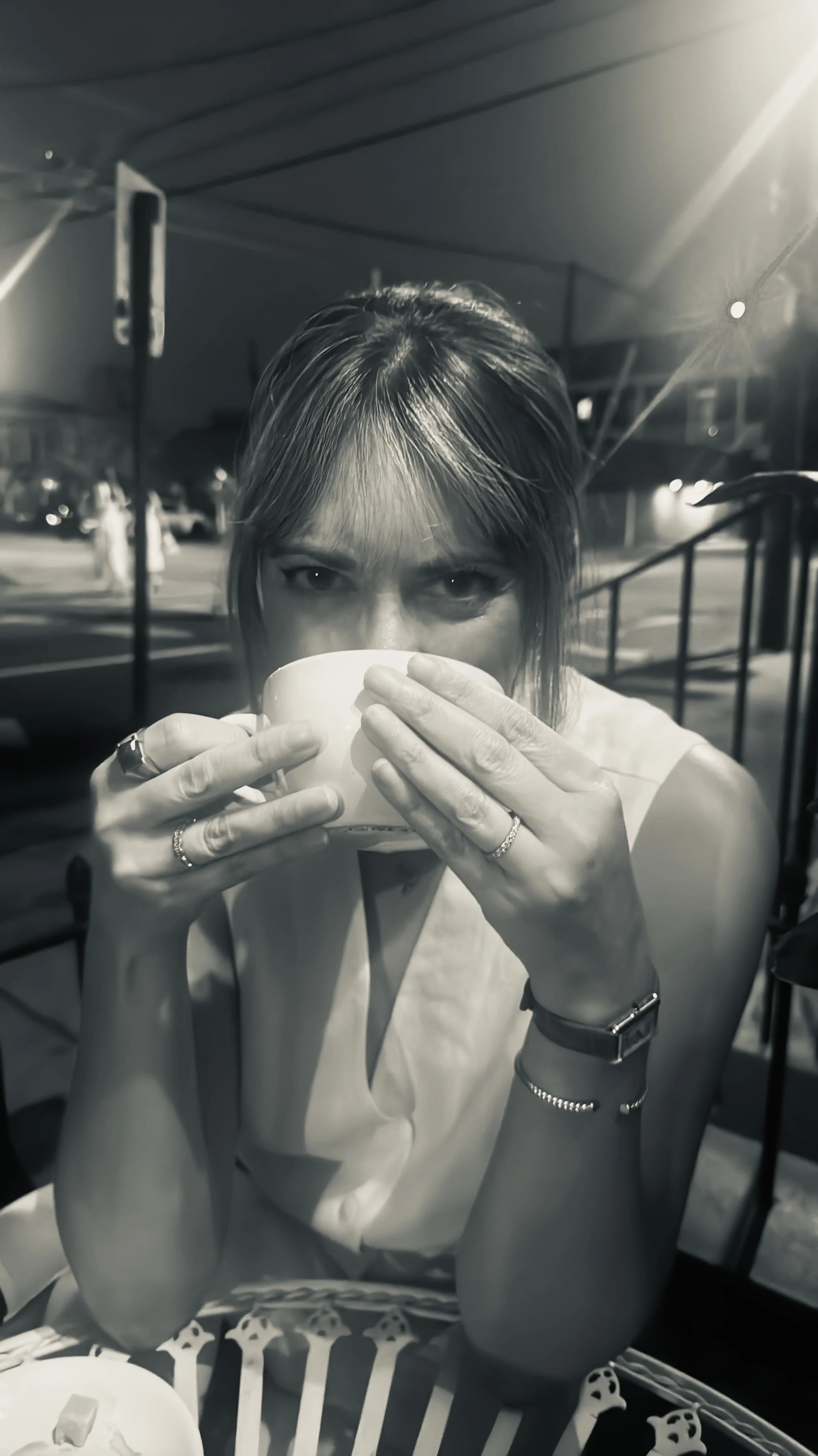 A woman with mid-length hair wearing rings and a watch is drinking from a large mug at an outdoor dining area during nighttime, with cars and streetlights in the background.