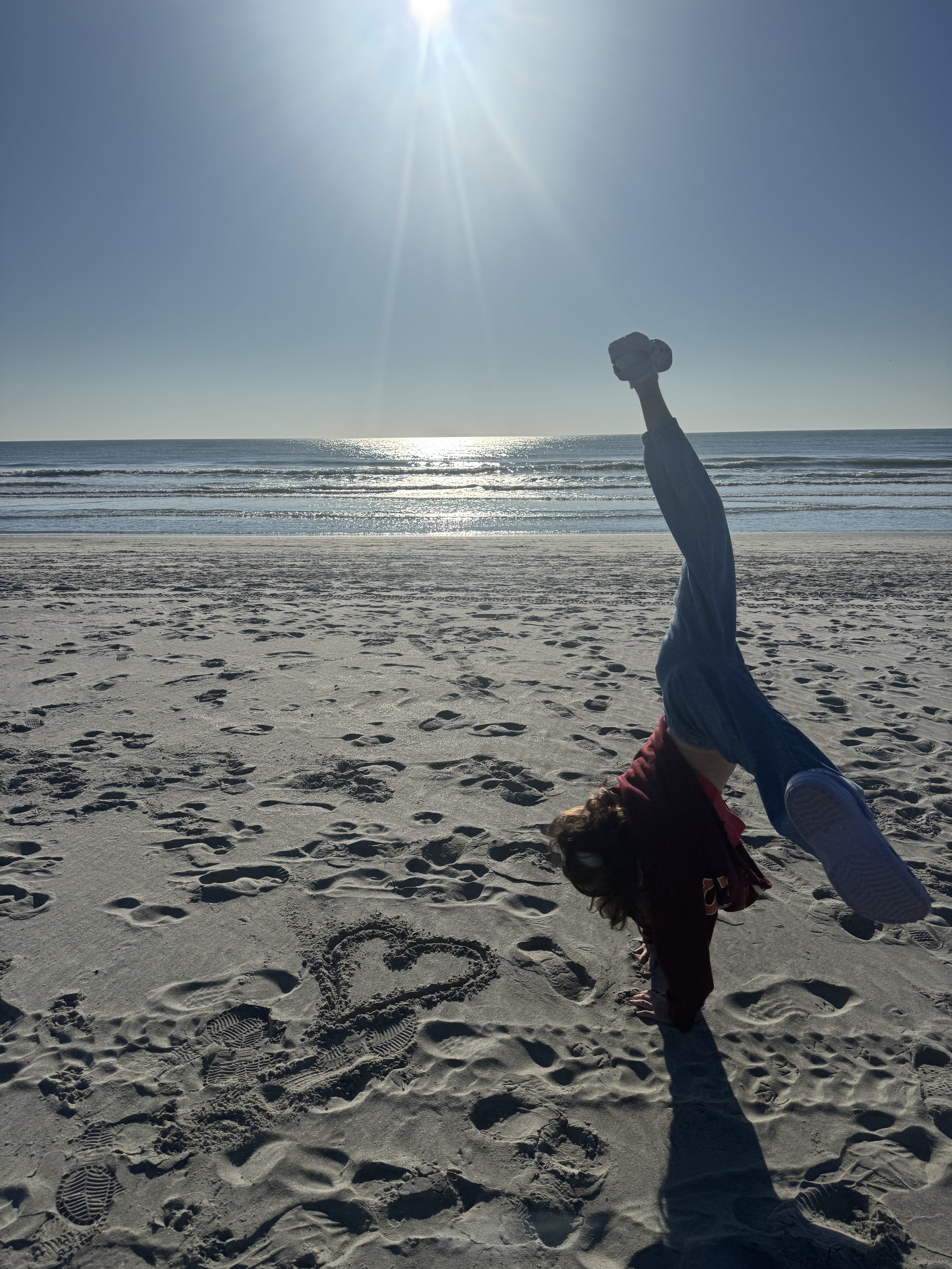 A person doing a handstand on the sandy beach near the ocean with the sun shining brightly overhead.