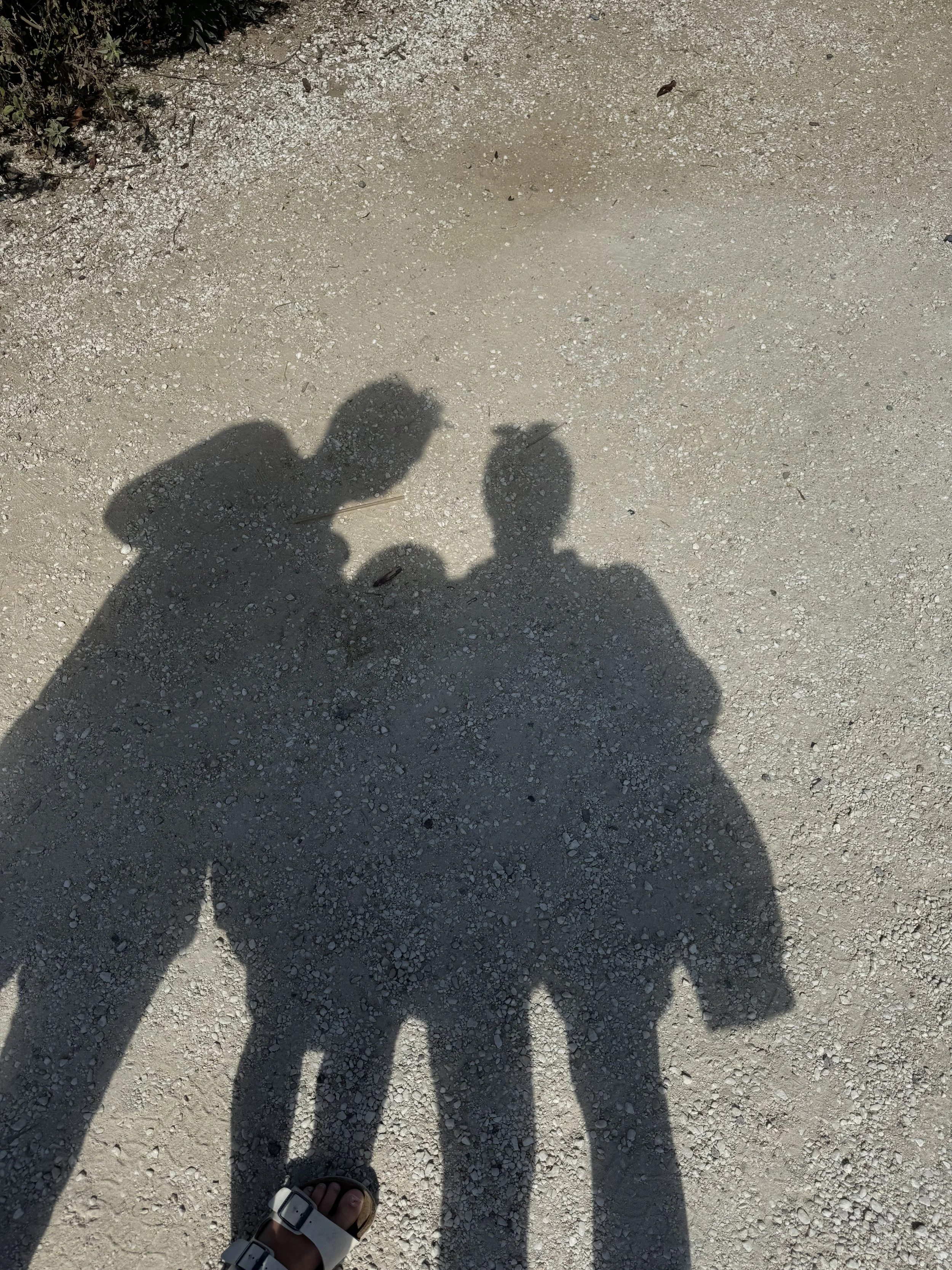 Shadows of two people and a child with a backpack on gravel ground.